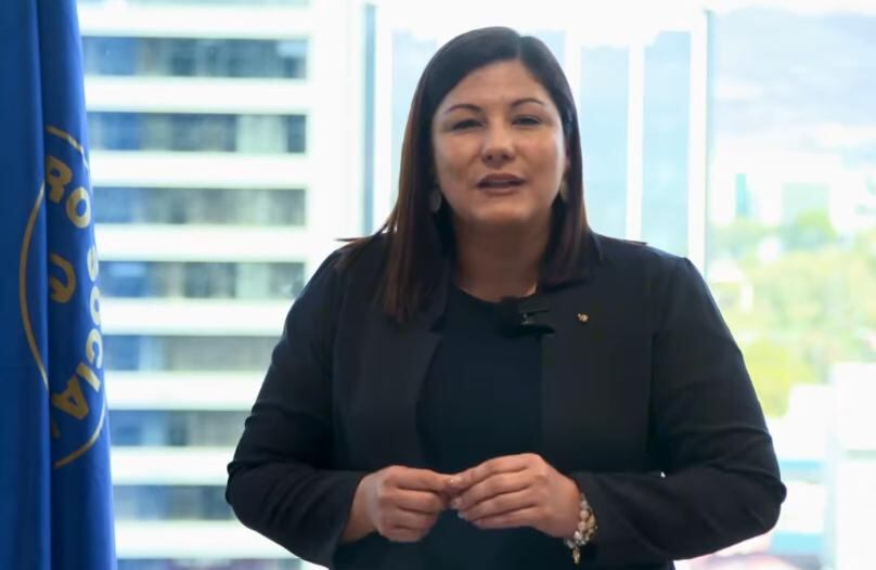 Fotografía de Mónica Taylor Hernández, presidenta de la Caja Costarricense de Seguro Social (CCSS), de pie frente a una ventana con vista a edificios y un fondo urbano. Lleva un traje formal oscuro y un micrófono de solapa, acompañada por una bandera institucional.