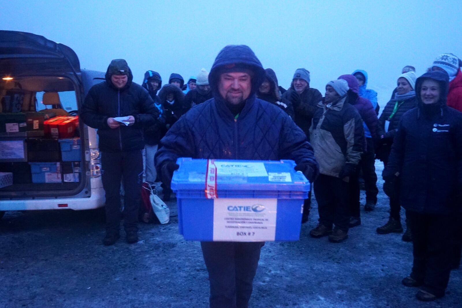 Un hombre carga una caja plástica en un ambiente de bajas temperaturas y nieve, atrás otras personas lo observan.
