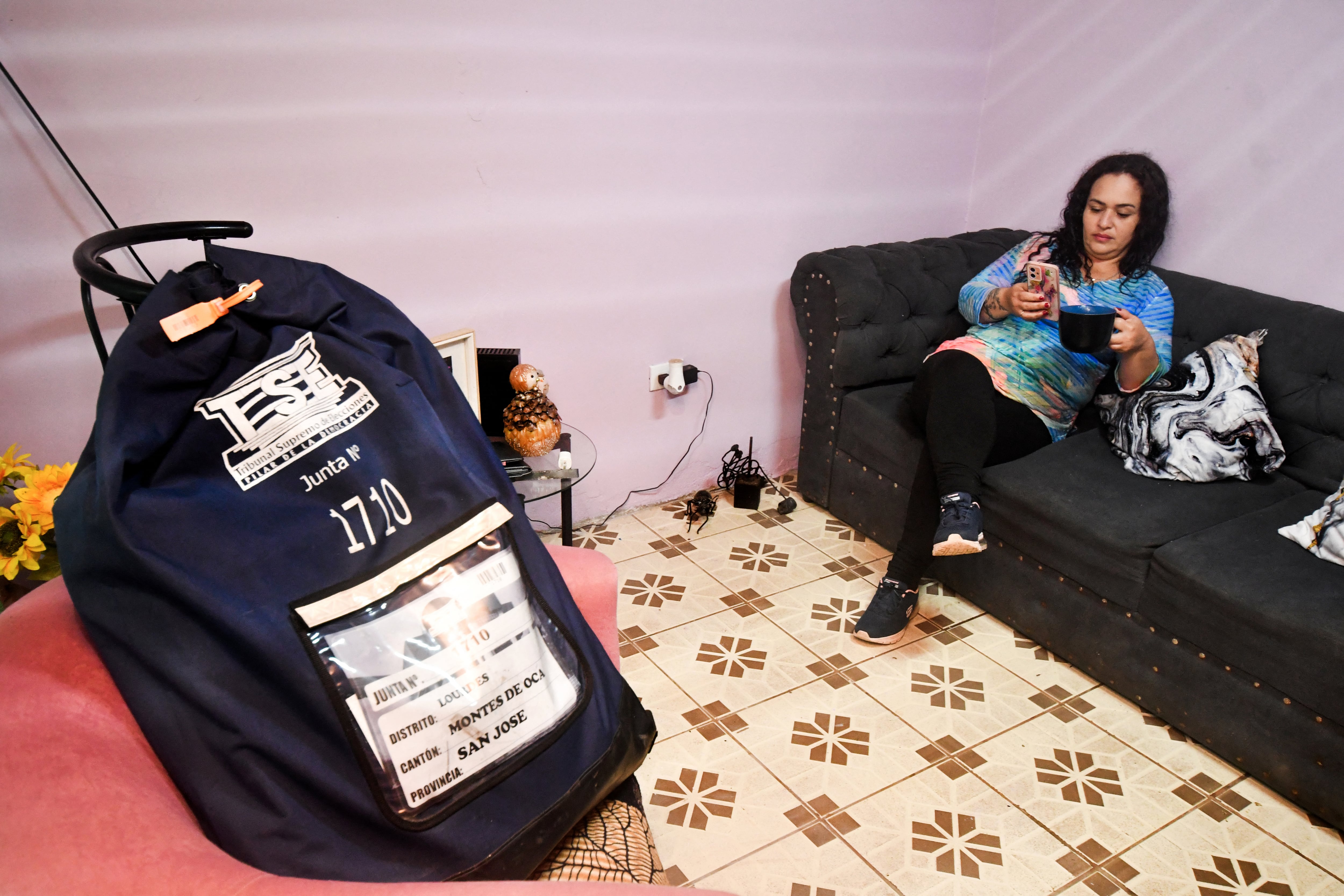 Priscilla Herrera, asistente de mesa electoral, toma café y revisa su teléfono móvil en su casa, junto a una bolsa que contiene material electoral, en San José, el 28 de enero de 2026. En Costa Rica, los miembros de mesa resguardan el material electoral en sus hogares hasta el día de las elecciones, cuando los votantes elegirán al próximo presidente y a los miembros de la Asamblea Legislativa el 1.º de febrero. (Foto de EZEQUIEL BECERRA / AFP)
