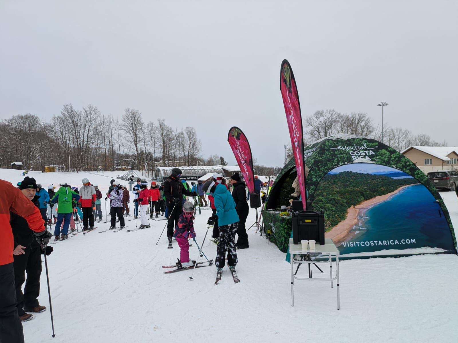 Personas caminando en un centro de esquí en medio de la nieve. Aparece una tienda de campaña con el nombre Costa Rica.