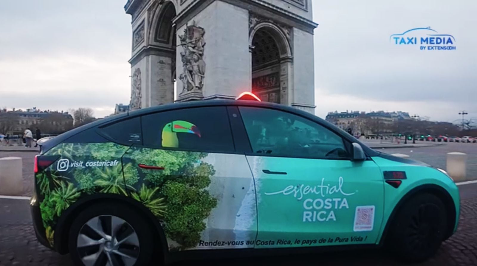 Taxi blanco transita por la rotonda del arco del Triunfo, en París, Francia. Luce un paisaje de bosque tropical con un tucán y el logo Esencial Costa rica. También un código de barras.