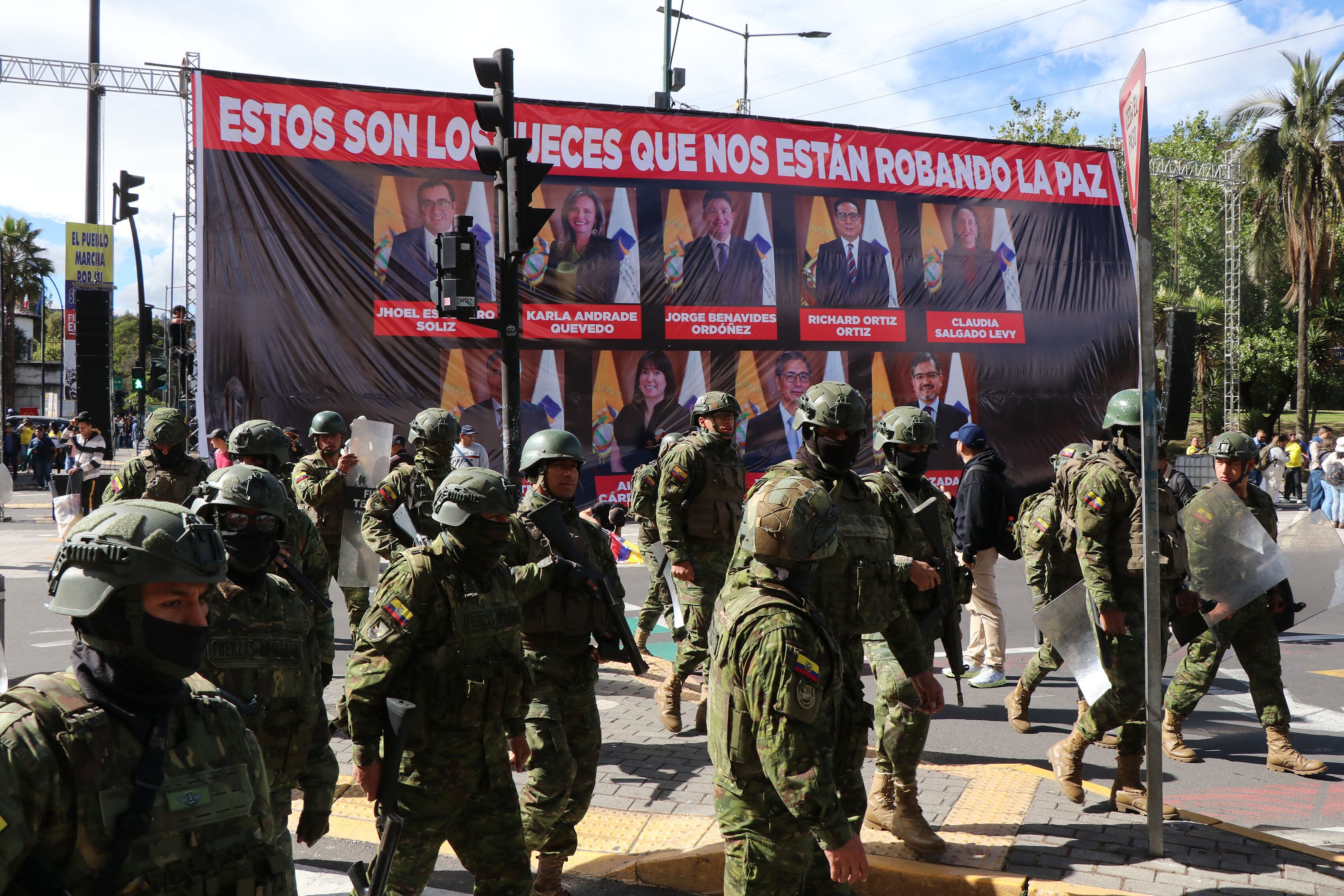Soldiers walk in front of a large banner with pictures of Constitutional Court judges, whom the government blames for insecurity in the country, displayed on Patria Avenue during a march called by Ecuador's President Daniel Noboa against the Constitutional Court's decision to censor several articles of recent laws in Quito on August 12, 2025. (Photo by Galo Paguay / AFP)