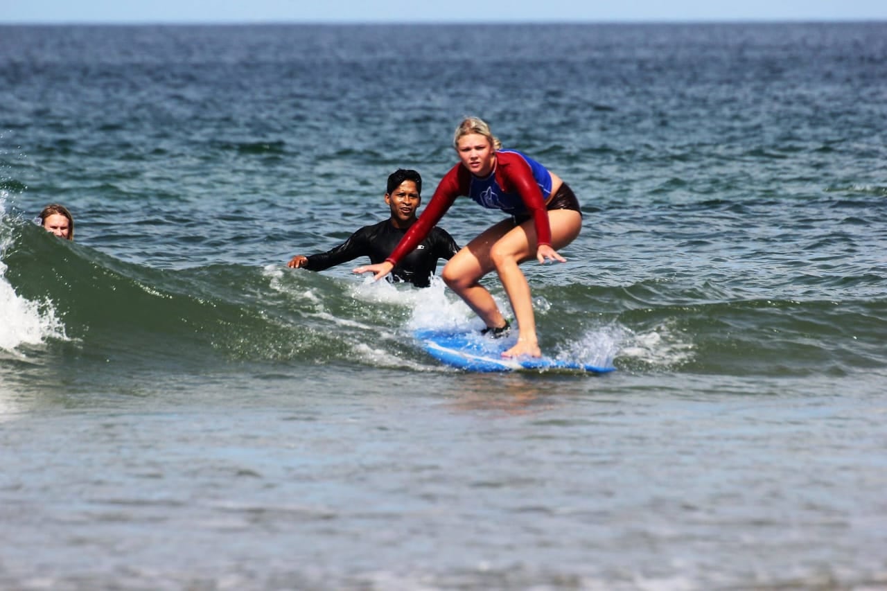 Turista sobre una tabla de surf montando una ola en una playa costarricense, con un instructor al fondo vigilando su progreso.