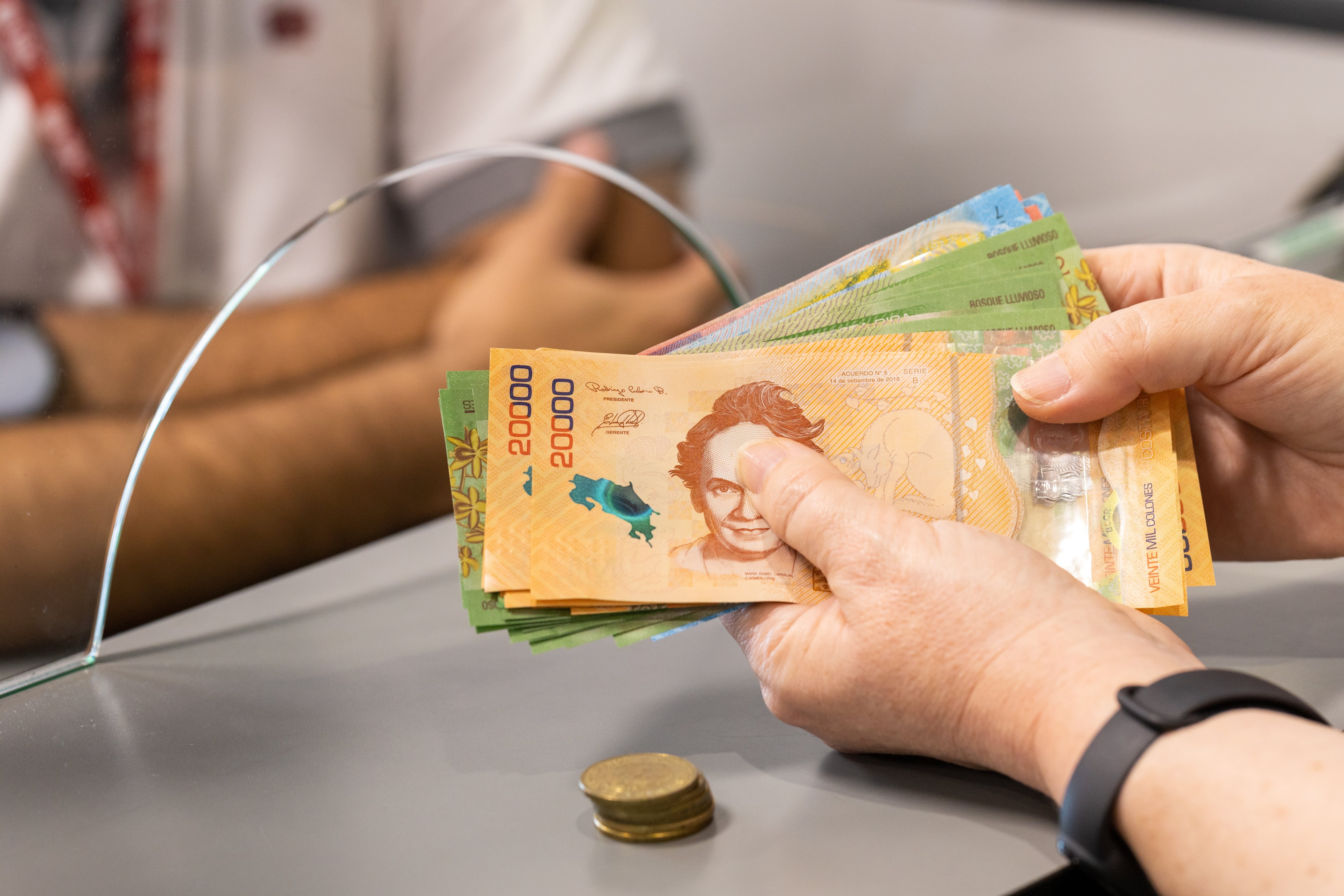 Una persona sostiene billetes de ¢20.000 y ¢10.000 en una ventanilla bancaria.