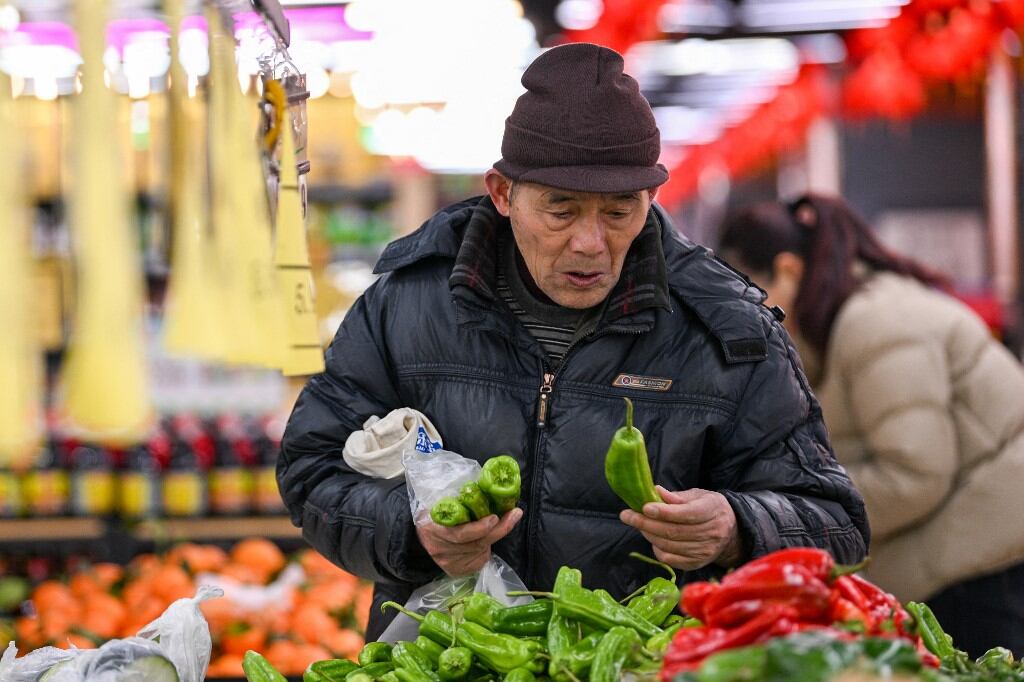 Hombre con chaqueta oscura y gorro selecciona pimientos verdes en un mercado en Pekín, China, rodeado de vegetales frescos.