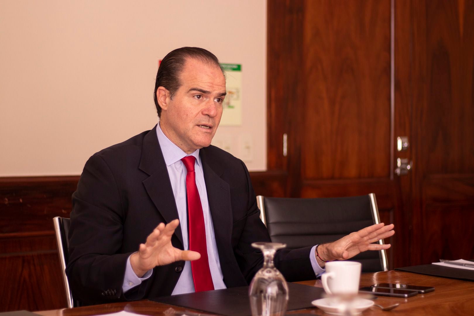 Hombre sentado de traje oscuro y corbata roja, gesticula durante una entrevista