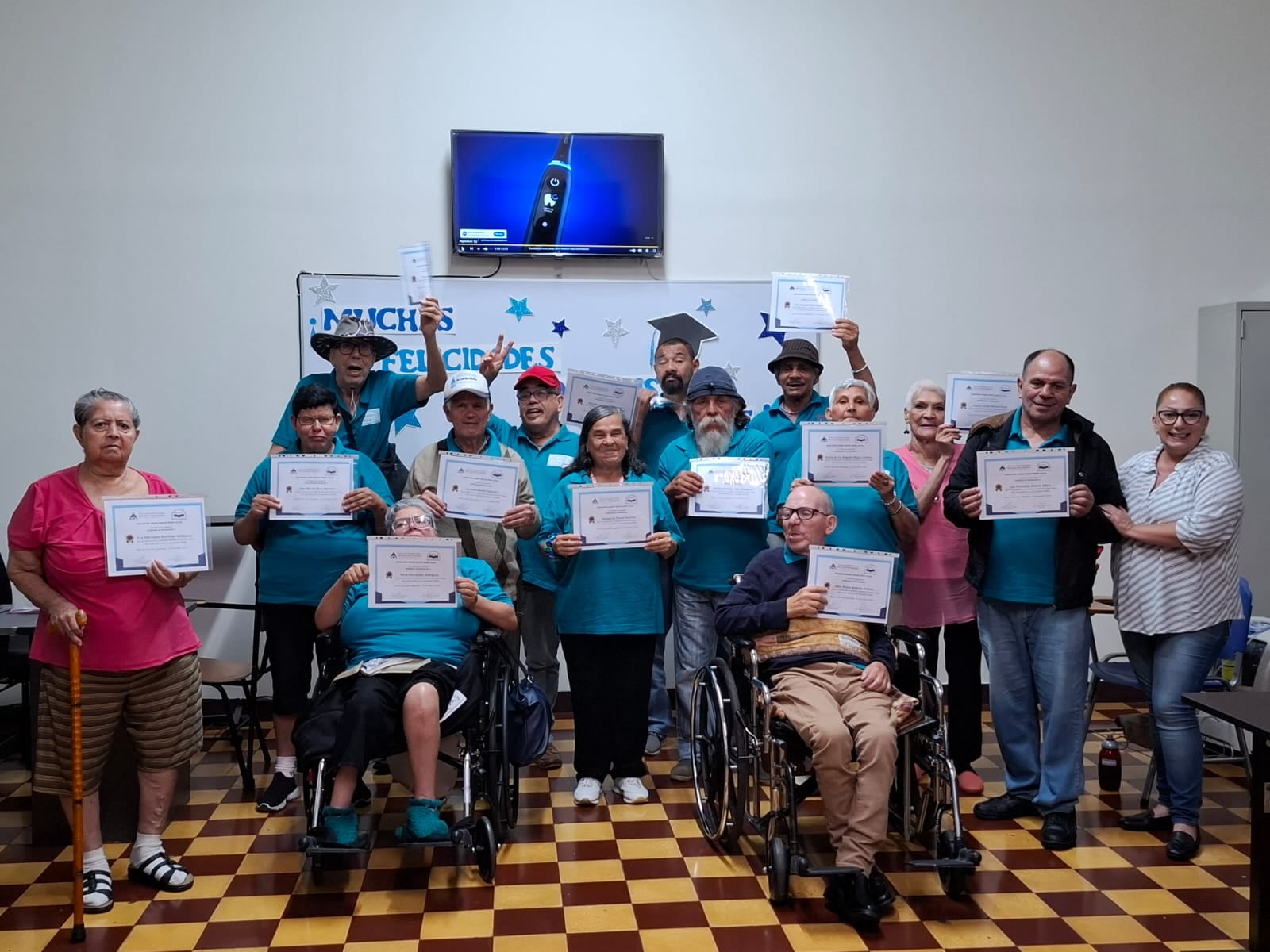 Retrato a grupo de 15 personas. Piso de cuadros amarillos y cafés. Pared blanca de fondo. Personas muestras título de graduación. Hay dos al frente en silla de ruedas. Todos son adultos mayores.