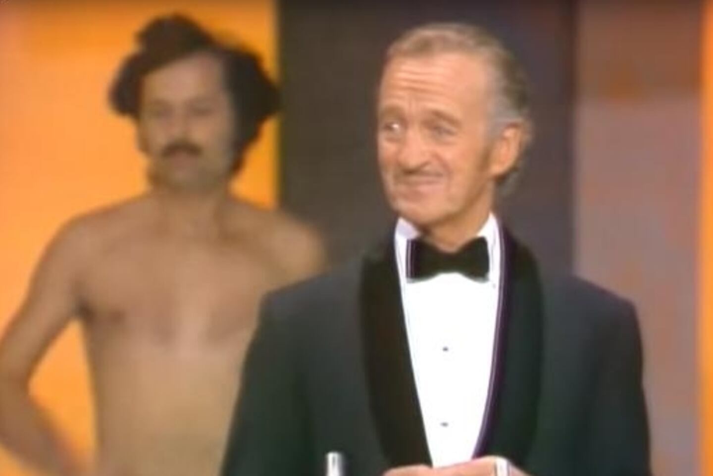 Presentador de los premios Óscar incómodo mientras detrás de él hay un hombre desnudo.