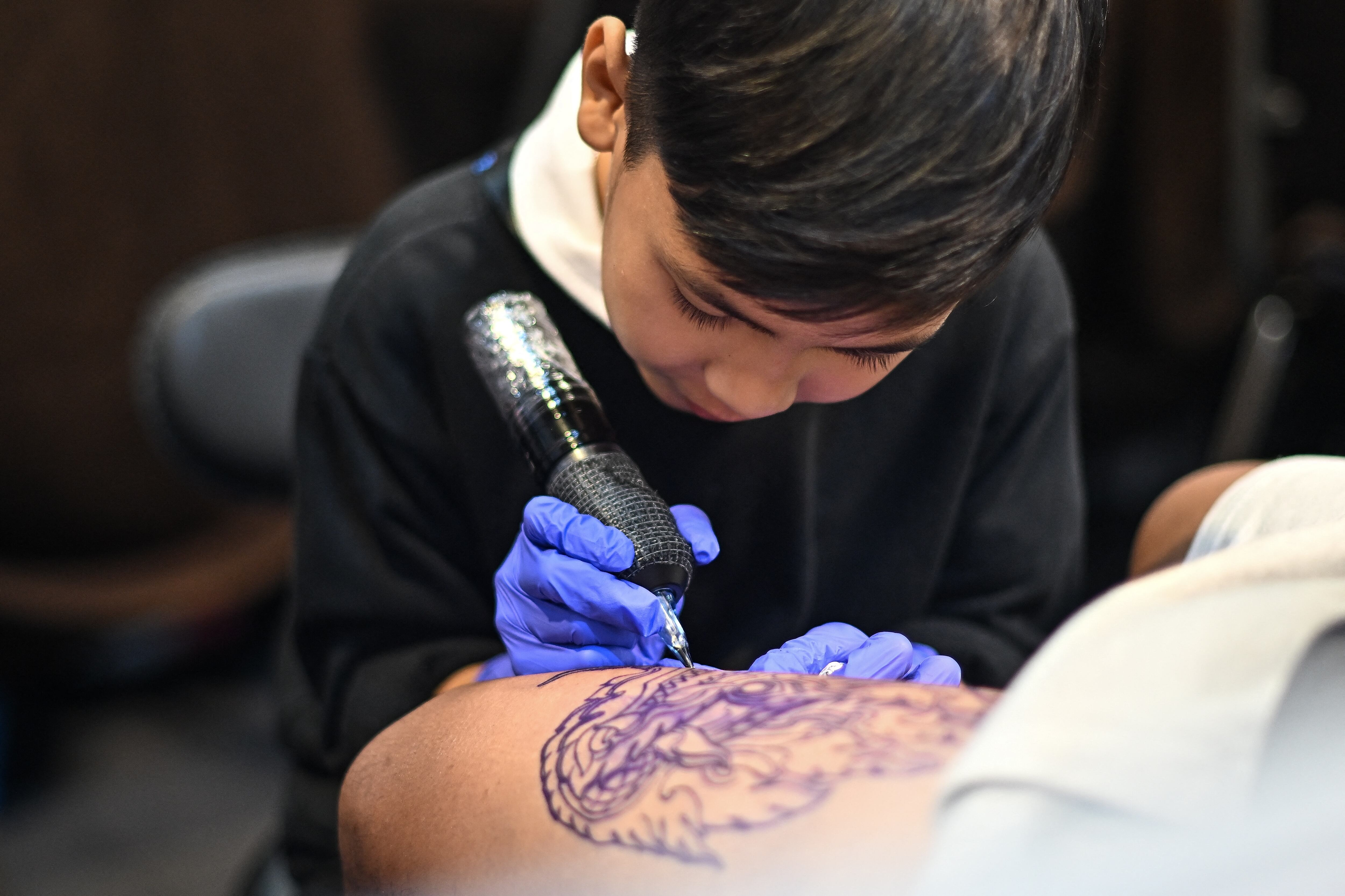 Nine-year-old Napat Mitmakorn, a Thai child tattoo artist works on his uncle�s tattoo at the 2025 Thailand Tattoo Expo in Bangkok on March 15, 2025. Wielding a cumbersome tattoo gun in child-sized hands swamped by adult-sized surgical gloves, nine-year-old Napat Mitmakorn expertly inks the pattern of a fanged serpent on a man's upper thigh. (Photo by MANAN VATSYAYANA / AFP) / To go with 'THAILAND-TATTO-CHILDREN-OFFBEAT' REPORTAGE by Montira RUNGJI