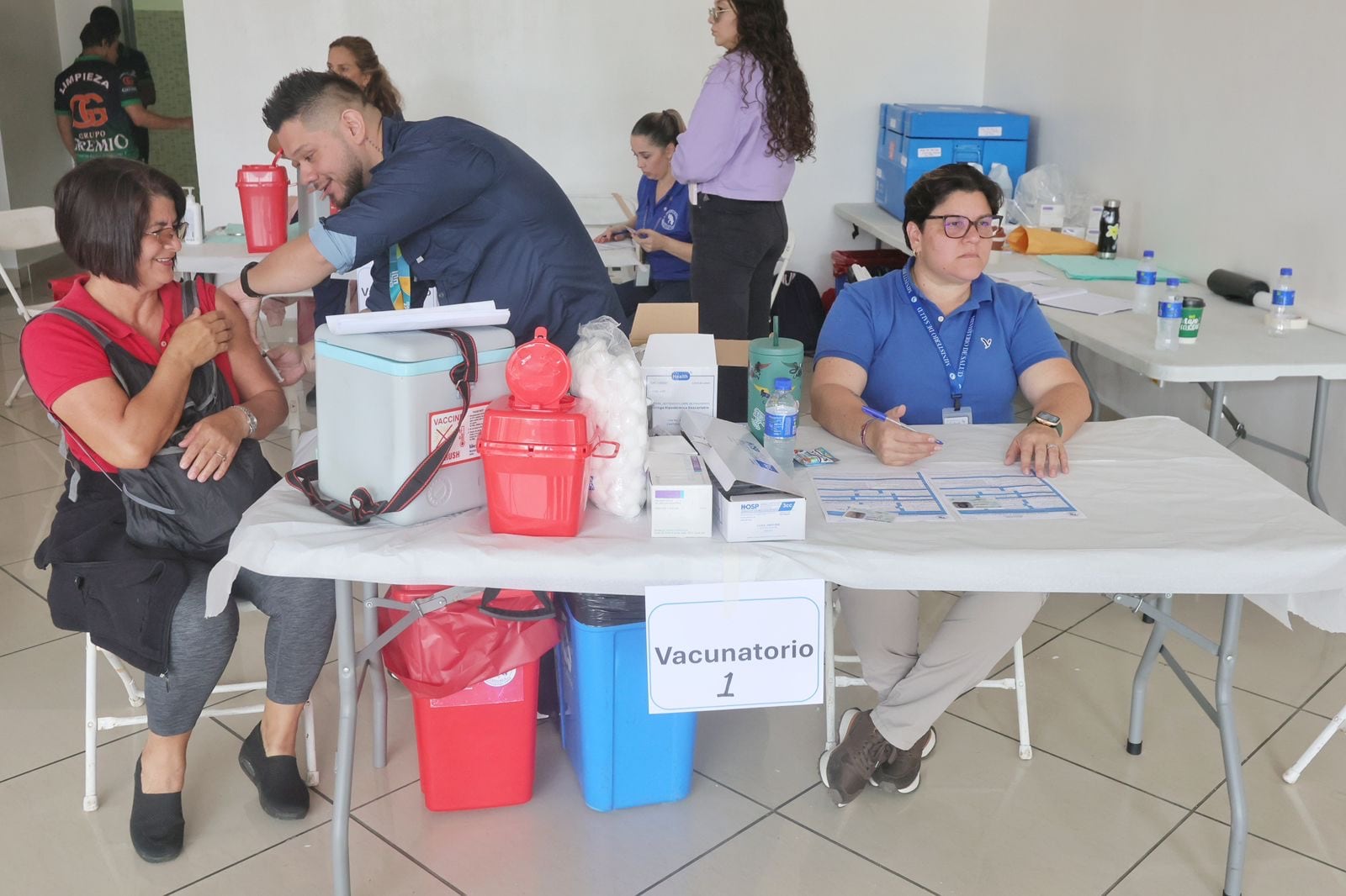 Tres personas alrededor de una mesa. Una de ellas, un hombre de camisa azul, con barba, inyecta a una mujer sentada en una silla, que viste blusa roja y pantalón gris. Es de pelo oscuro. Otra mujer de pelo corto, negro, está sentada a la mesa, que está con implementos para vacunar, como hieleras para vacunas.