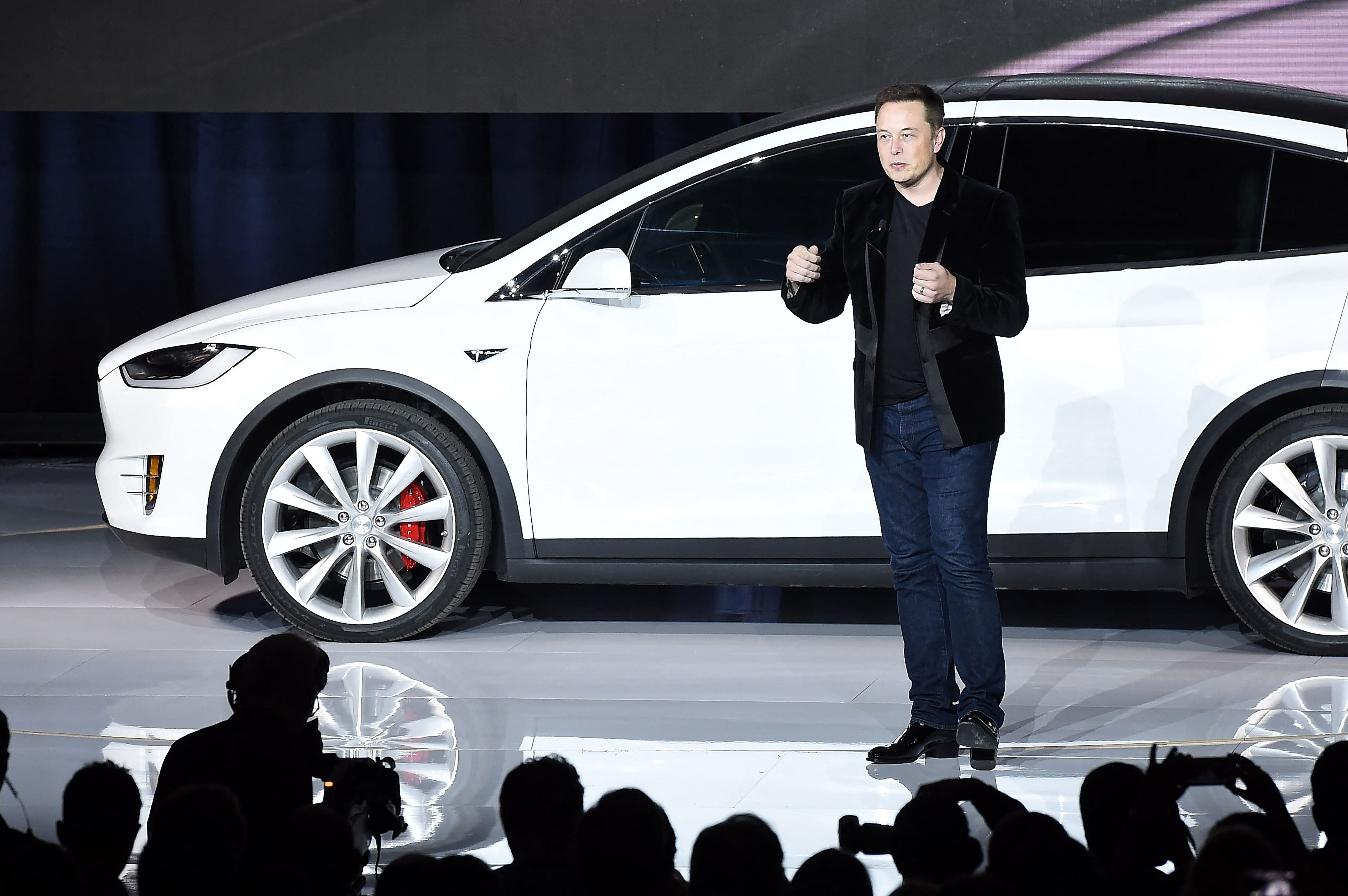 Elon Musk, vestido de traje negro, de pie junto a un Tesla Model X blanco, en un evento de presentación oficial.