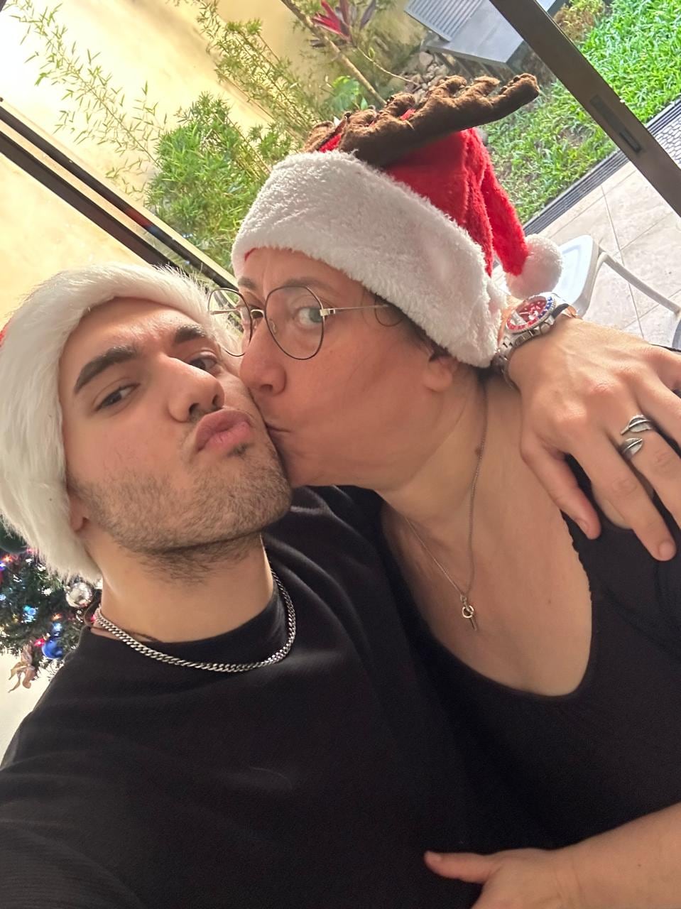 Un joven recibe un beso en la mejilla de su madre; ambos posan con sombreros navideños