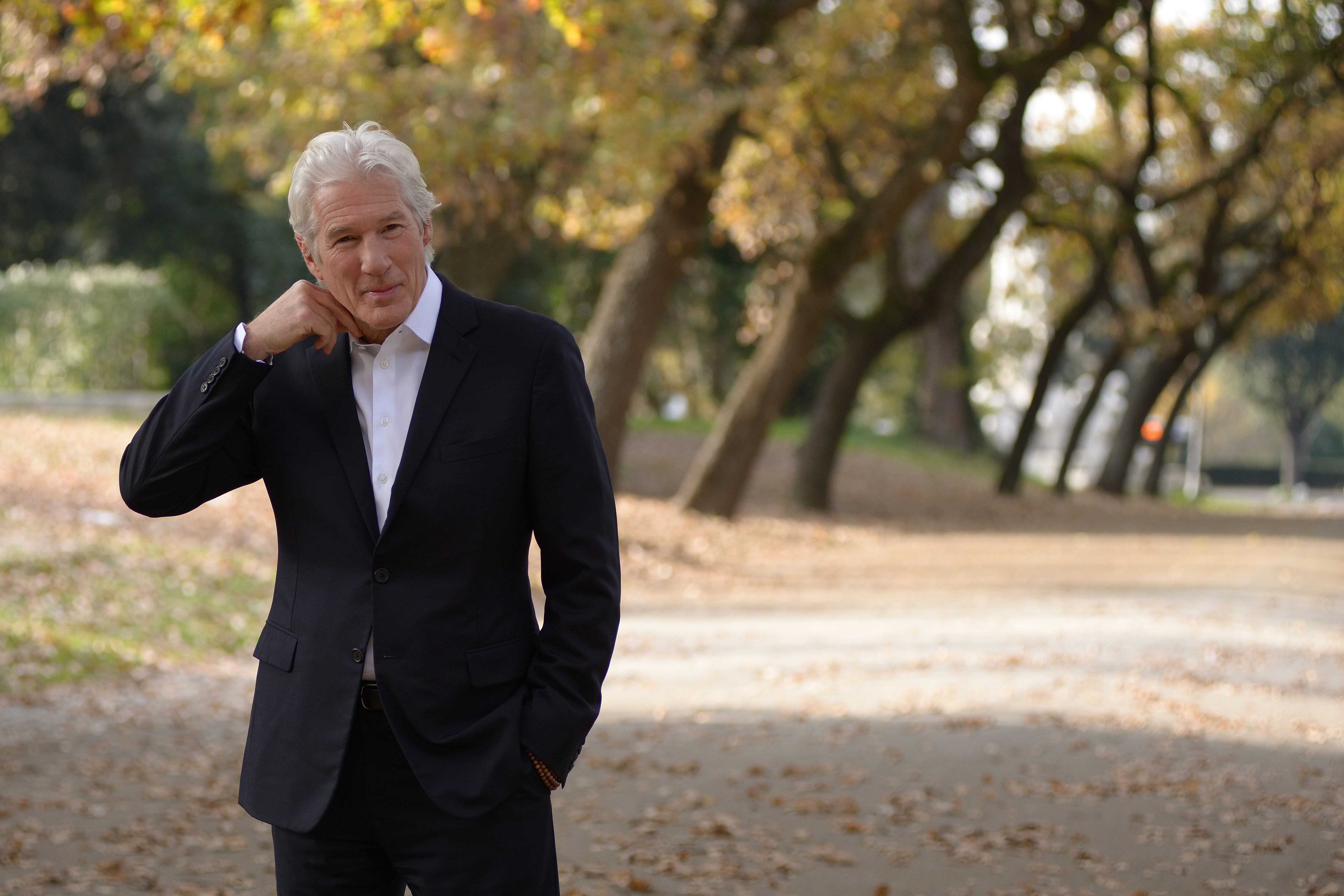 Richard Gere posando para la foto mientras camina. Usa traje negro y camisa blanca.