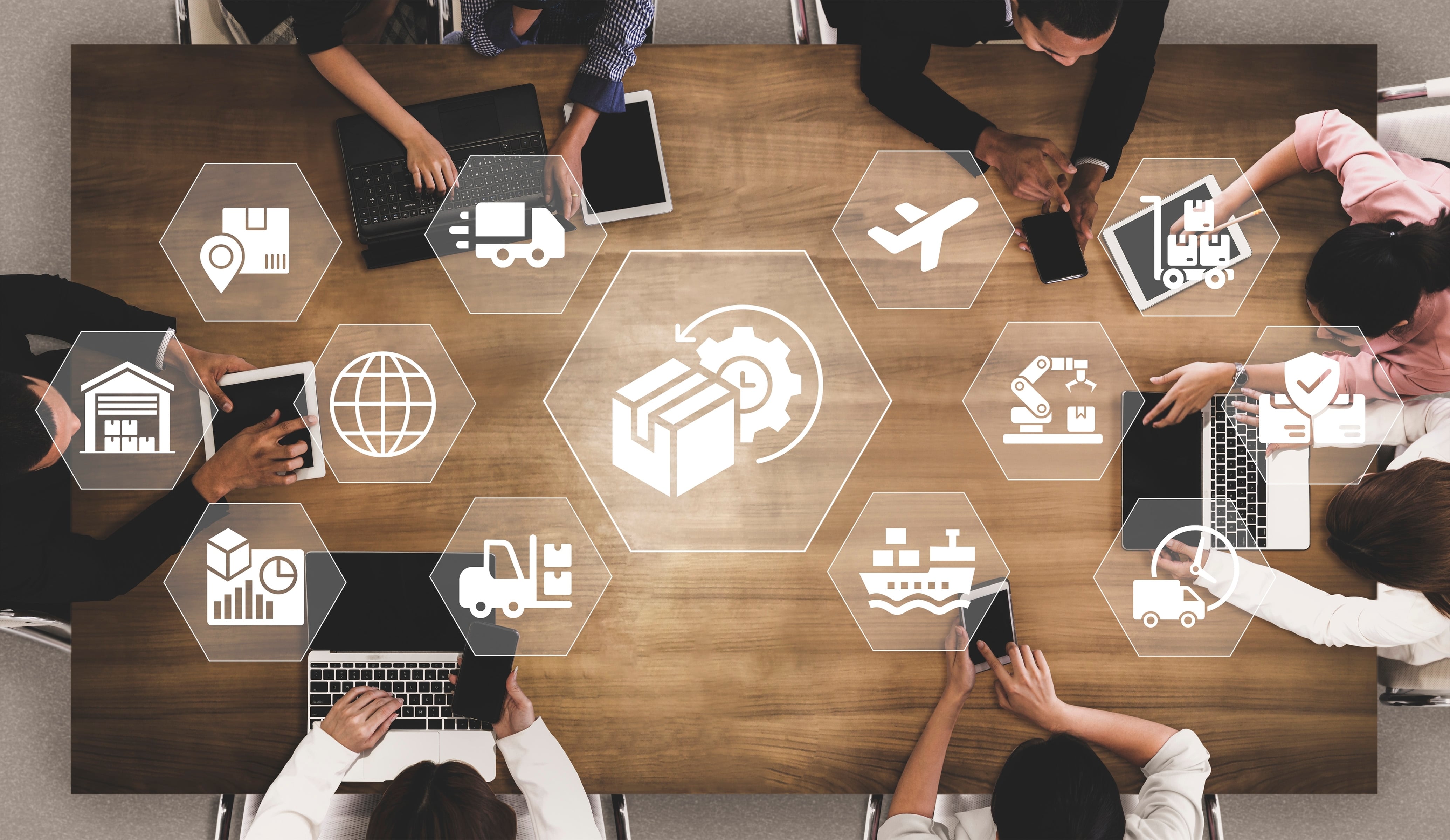 Vista aérea de un grupo de personas reunidas alrededor de una mesa de trabajo utilizando computadoras, tabletas y teléfonos móviles. Sobre la imagen aparecen iconos digitales relacionados con logística, transporte, almacenamiento, manufactura, comercio internacional, distribución y cadenas de suministro. La composición representa procesos de comercio exterior e integración económica.