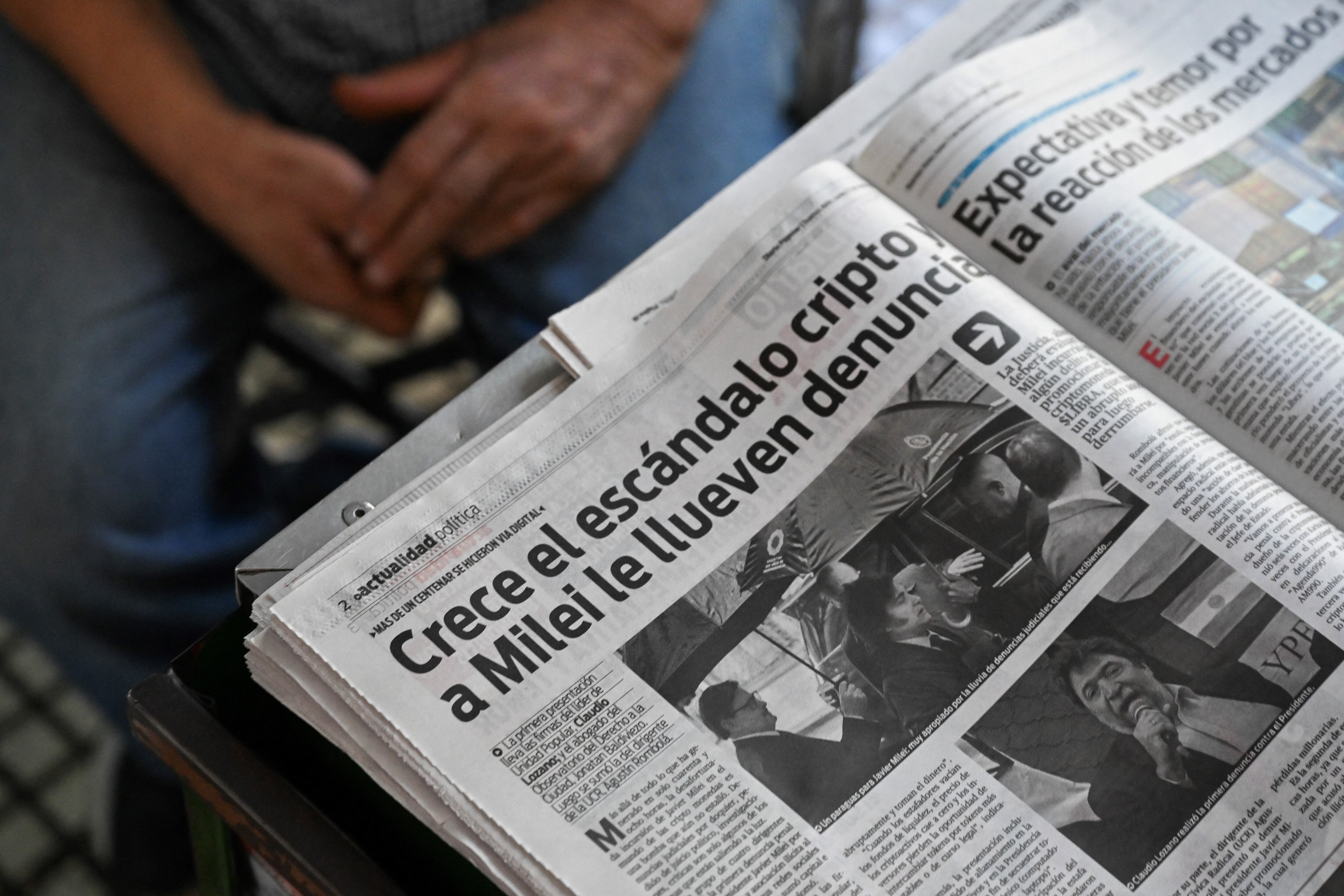 Primer plano de un periódico con la noticia sobre el escándalo cripto de Javier Milei, con el titular en negrita y fotografías relacionadas. Un hombre sentado observa la publicación.