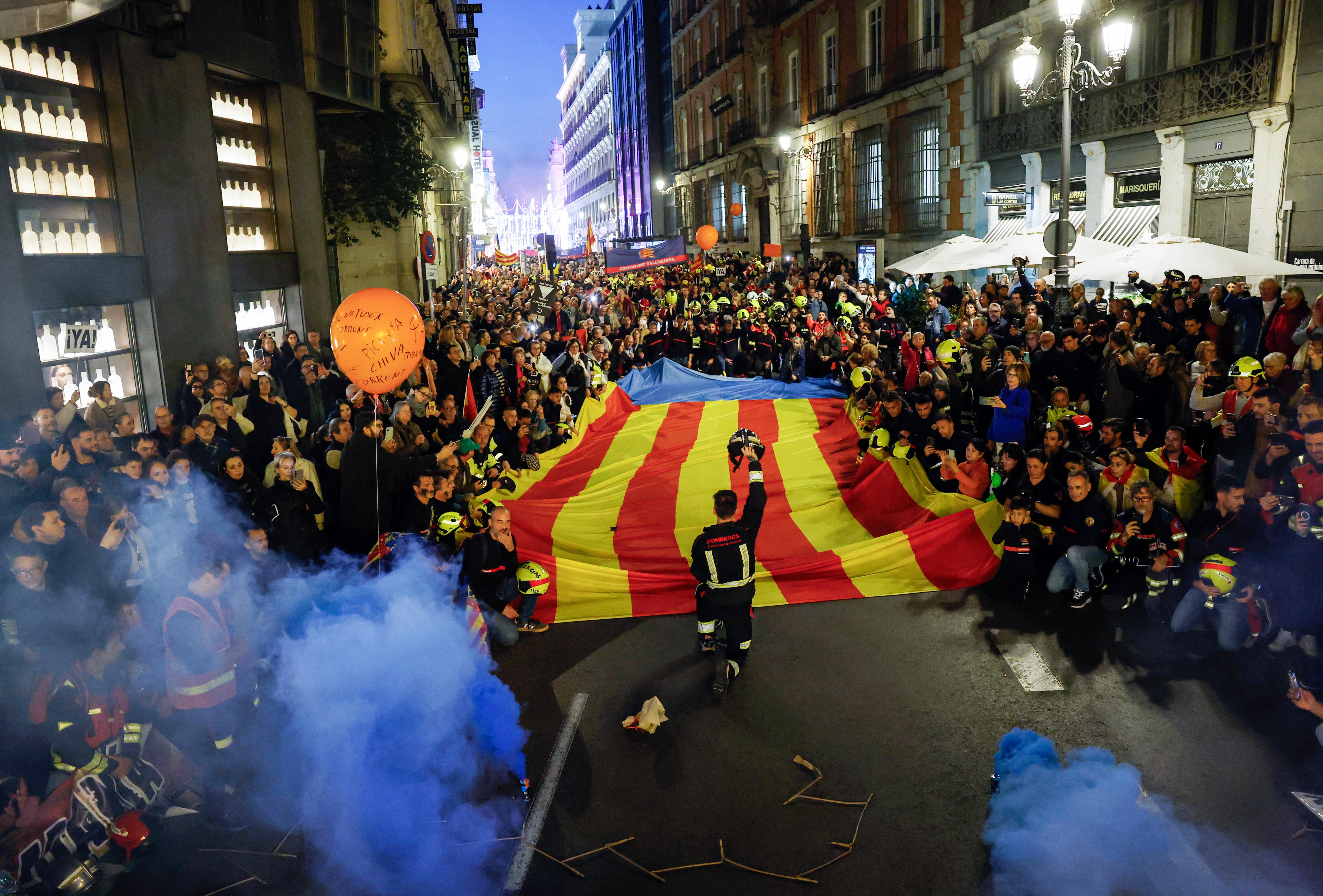 Bomberos en protesta sosteniendo una bandera gigante de Valencia, exigiendo respuestas por la falta de coordinación en la gestión de inundaciones en la región de Valencia, Madrid.