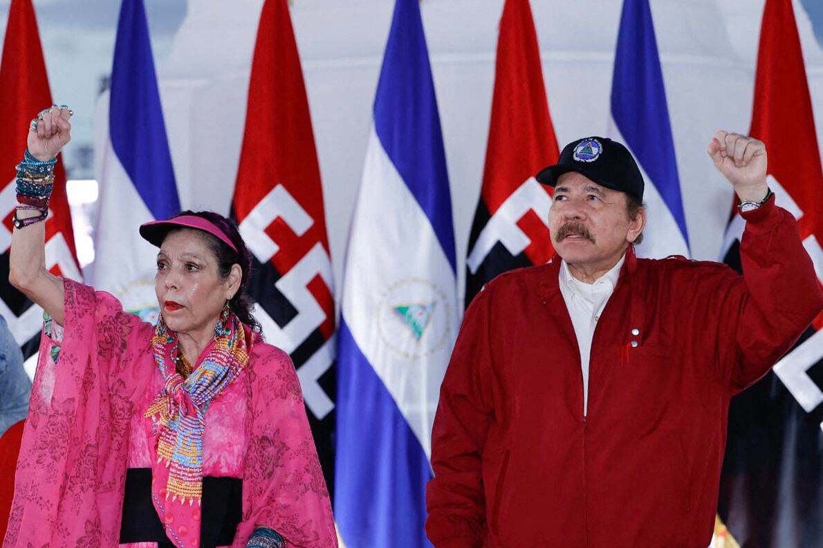Daniel Ortega y Rosario Murillo, vestidos con ropa roja y rosa respectivamente, levantan el puño en un evento oficial en Nicaragua, con banderas nacionales y del Frente Sandinista de Liberación Nacional (FSLN) de fondo.