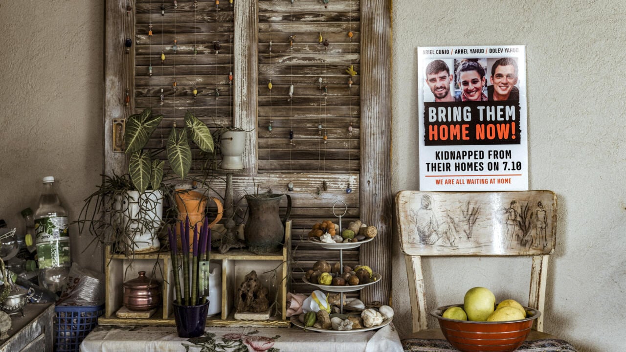 Cartel con fotografías de Arbel Yehud y otros rehenes, con el mensaje "Bring them home now", rodeado de plantas y objetos decorativos en un espacio interior.