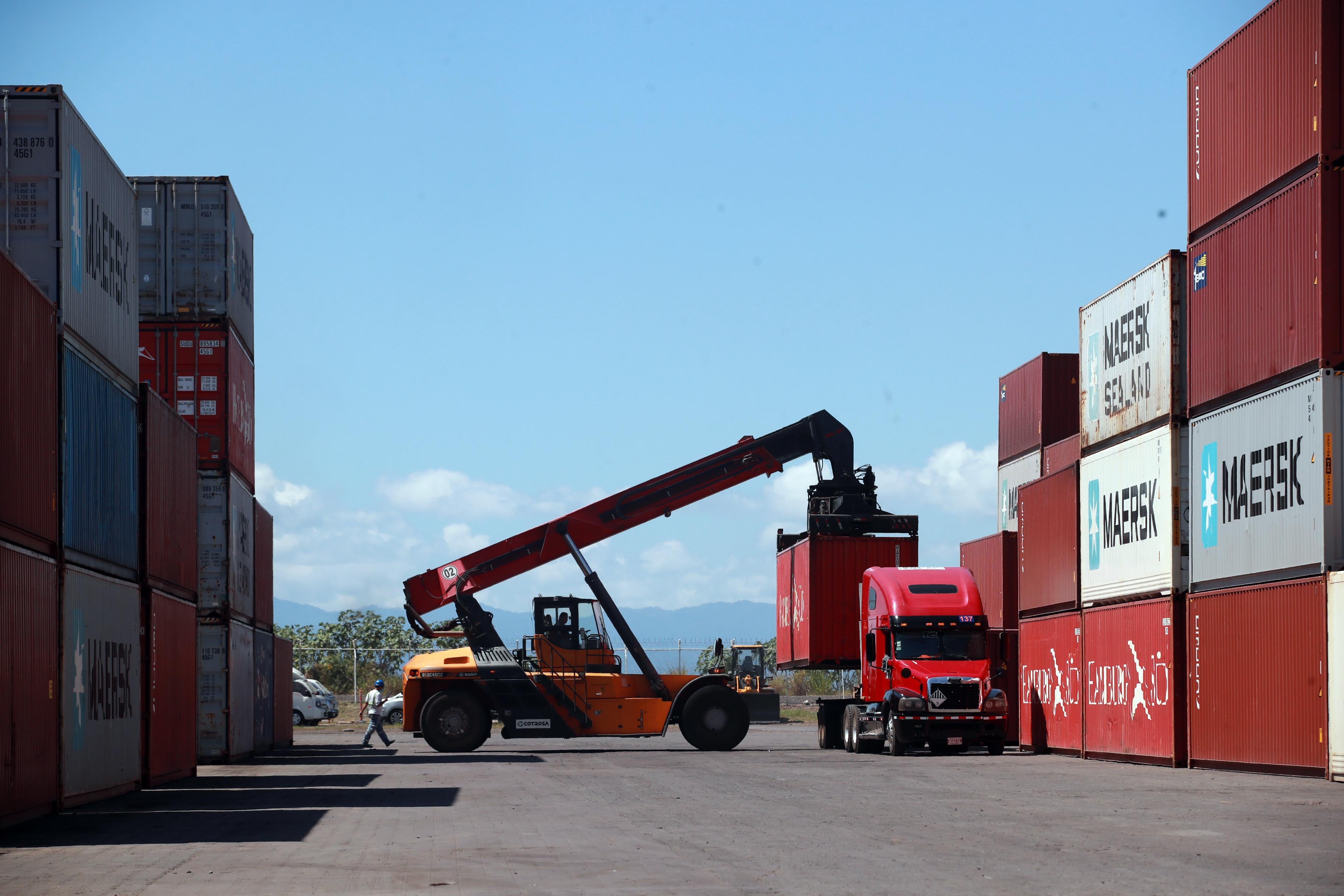 Montacargas moviendo un contenedor en una terminal de carga, rodeado de camiones y contenedores, bajo un cielo despejado.