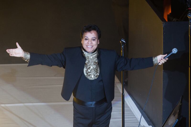 Juan Gabriel en un concierto, con sus brazos extendidos en señal de abrazo y una gran sonrisa. Usa un traje y camisa negros.