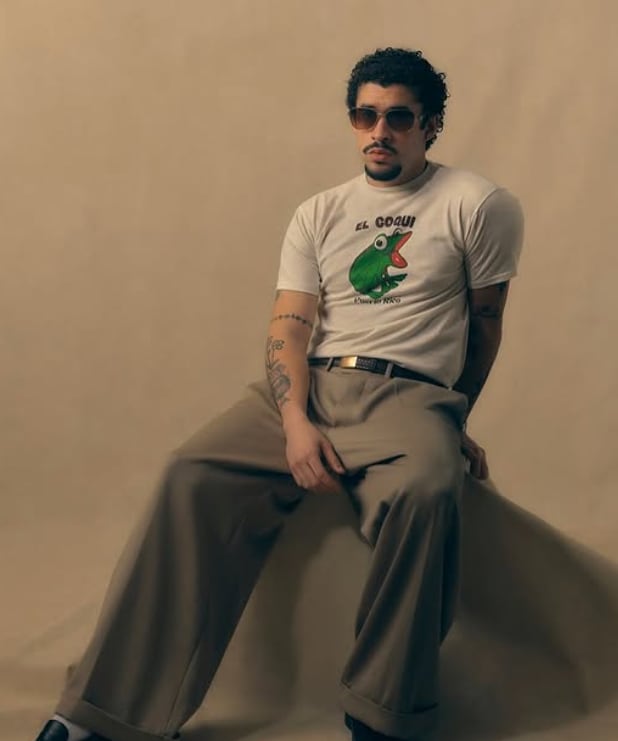 Imagen del puertorriqueño Bad Bunny sentado. Usa lentes de sol, una camiseta con un dibujo de un ave verde.