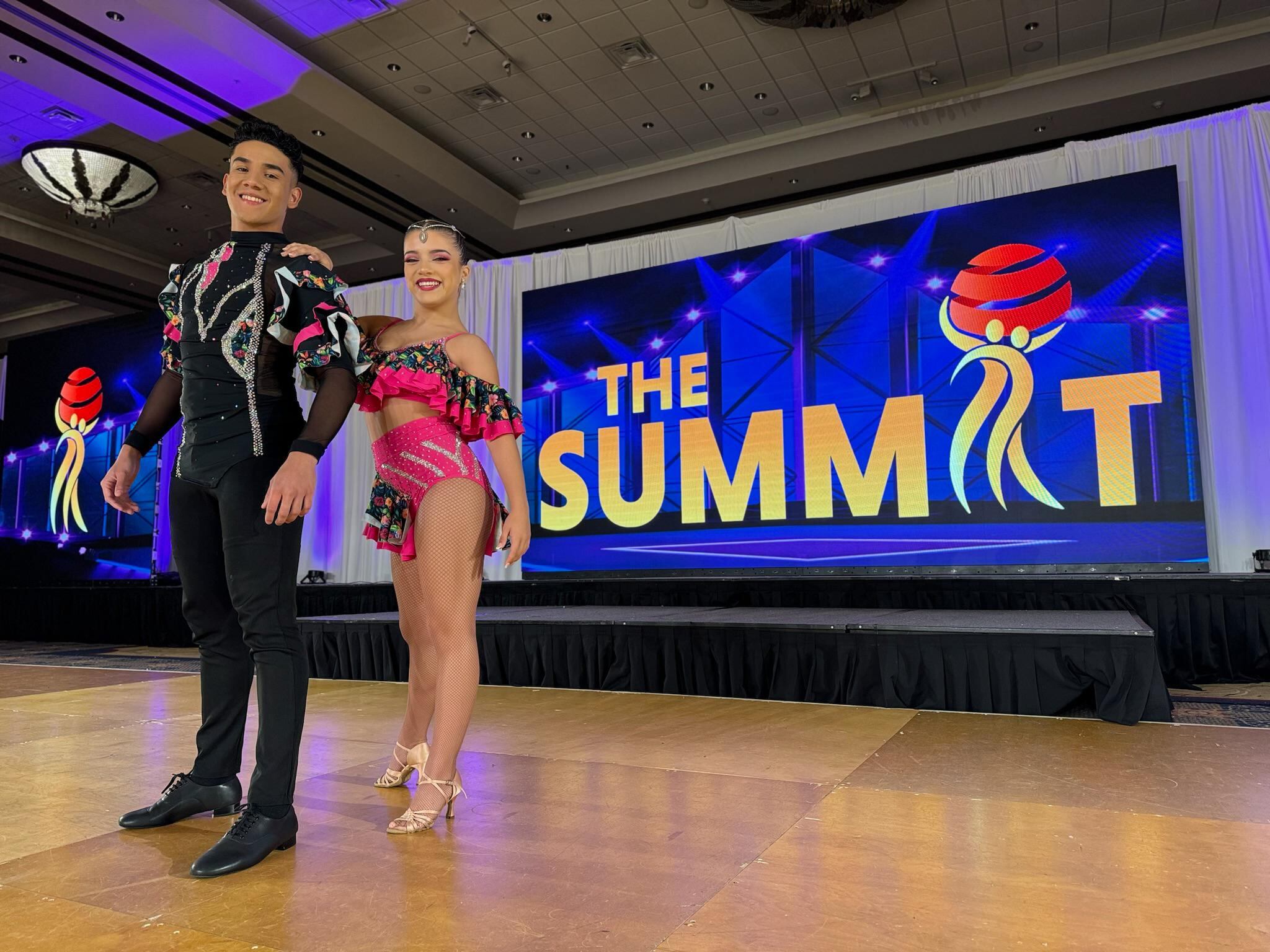Imagen de la bailarina costarricense Mariangel Gomez junto a su compañero de baile Allan Sánchez. Posan con sus trajes de competencia de baile, él de negro y ella con un vestido de tono rosado.
