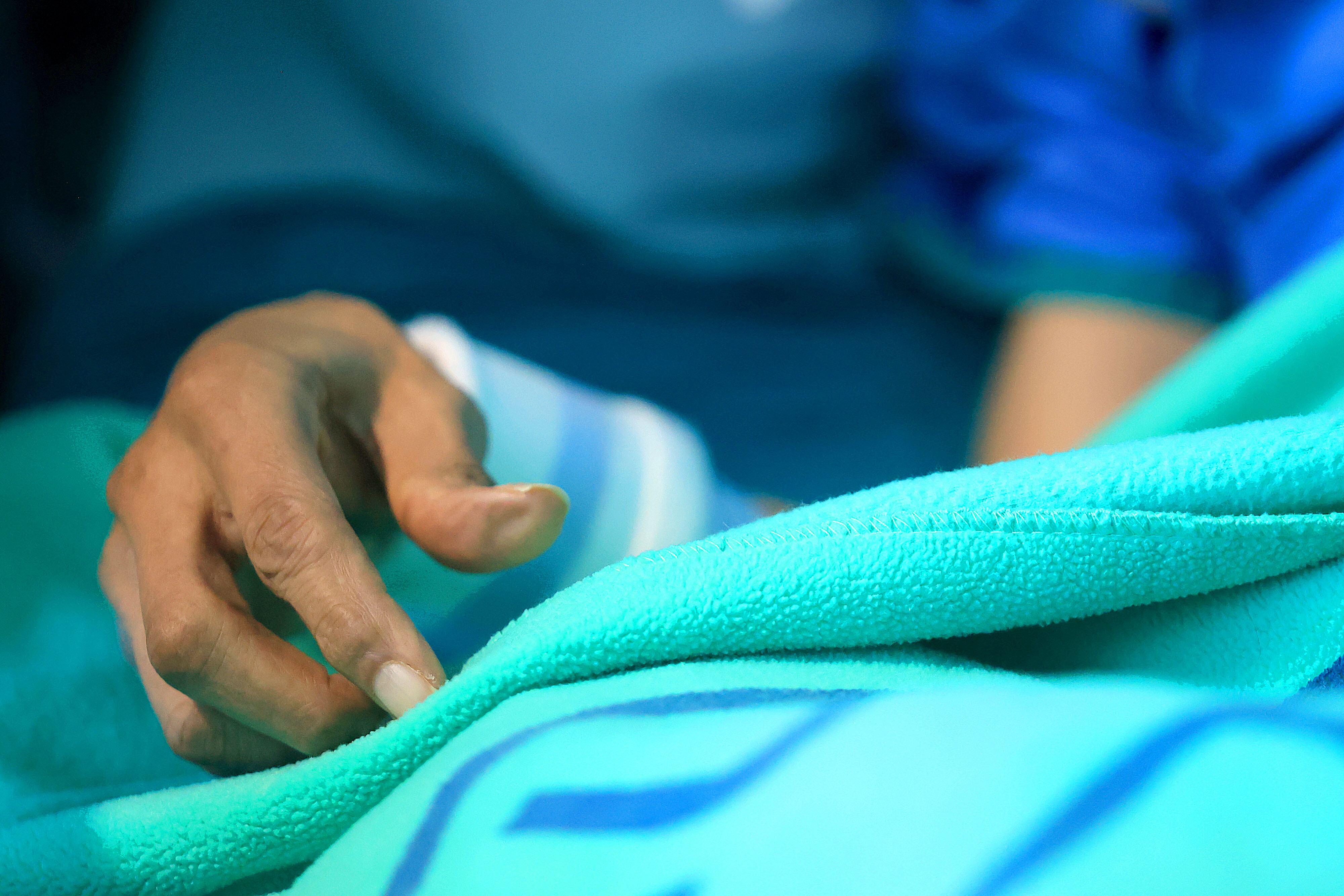 Mano de un adulto mayor, delgada, sobre una cobija. Al fondo, parte del cuerpo de la persona.