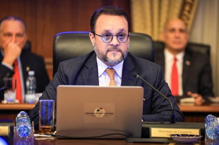 Hombre de traje oscuro y corbata naranka con lentes sentado con una computadora portátil. Al fondo se observan otras personas desenfocadas en una sala de conferencias.