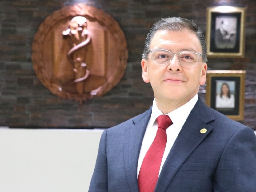Retrato de hombre de mediana edad, blanco, delgado, con traje entero azul, corbata roja, manos enlazadas al frente.