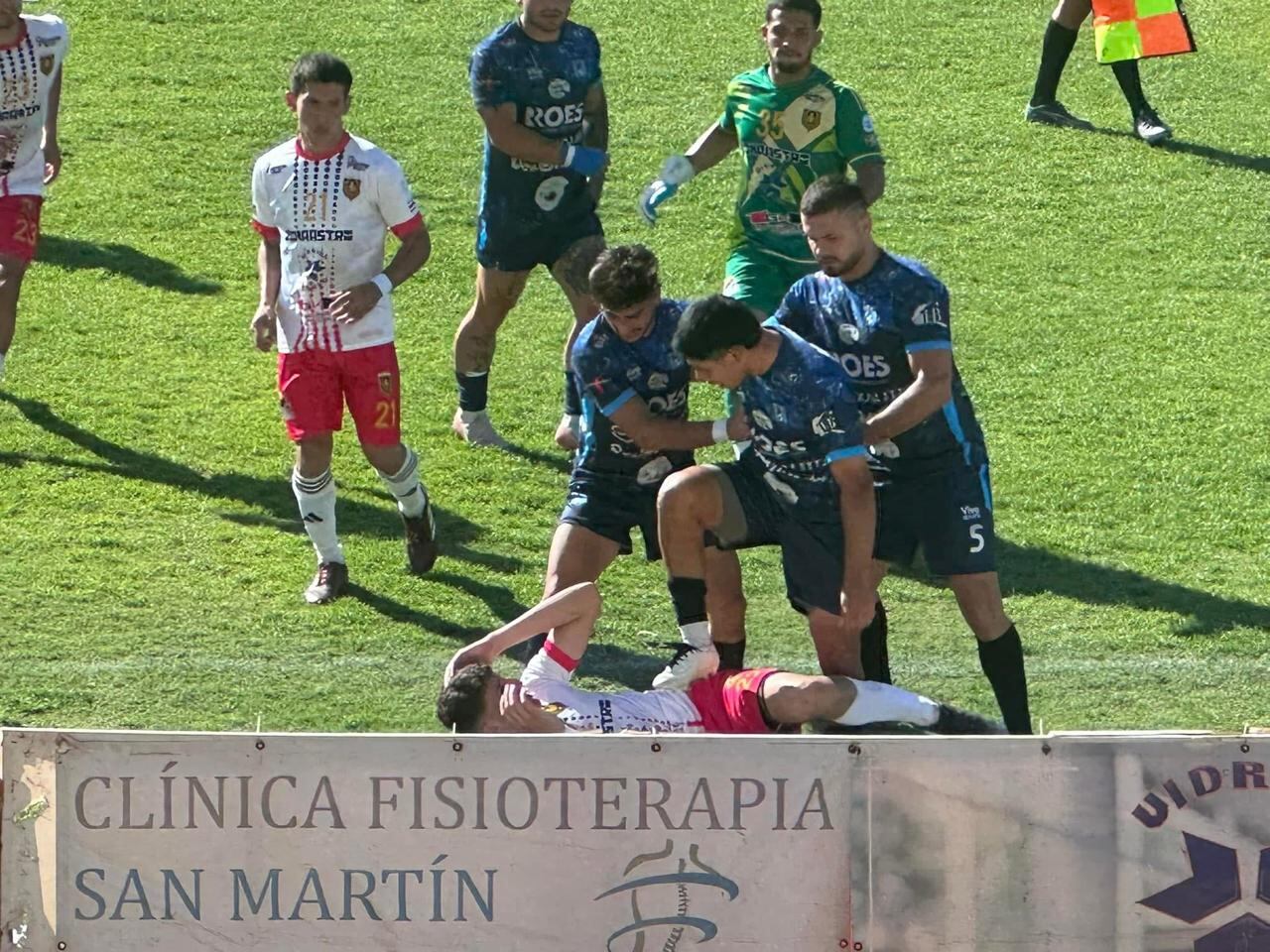 Un jugador con el uniforme azul de Rosario de Naranjo pisotea a uno con uniforme blanco de la Águilas de Liberia, rodeados por futbolistas de ambos equipos.