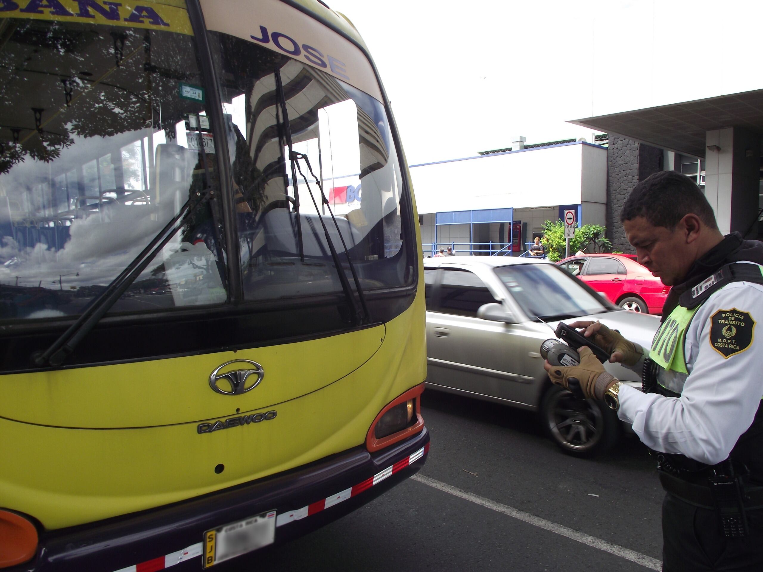 Una persona con uniforme de oficial de Tránsito parado frente a un bus, apuntando en una libreta, como haciendo una multa.