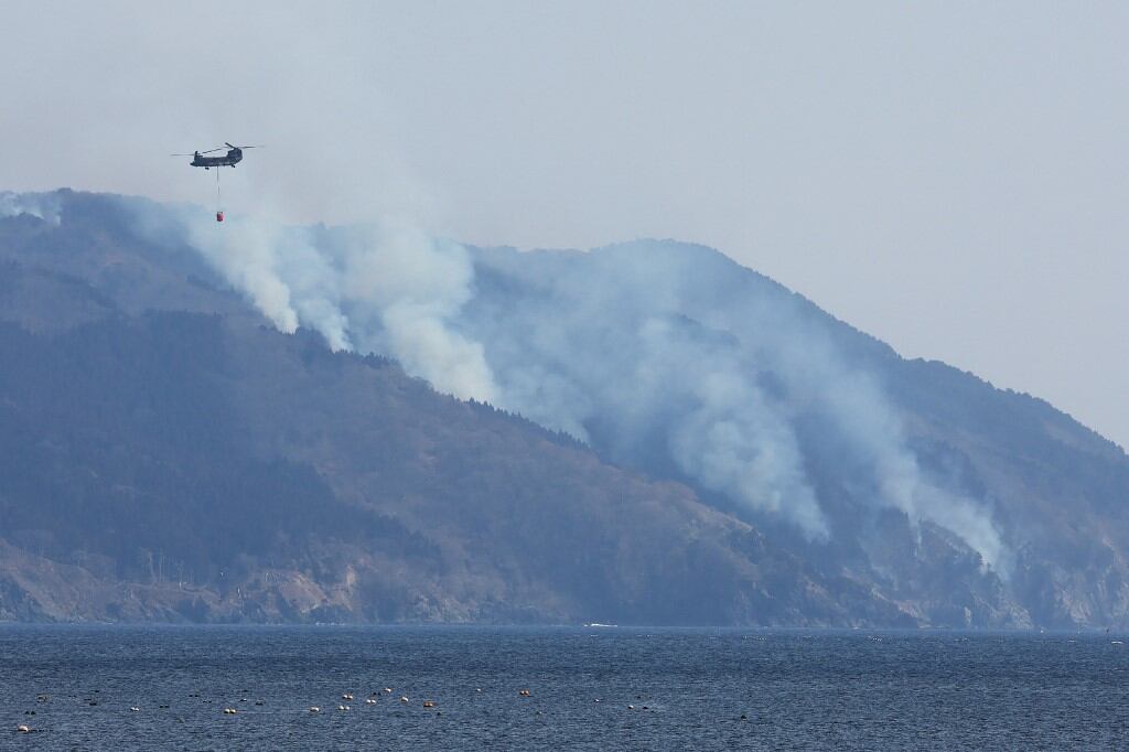 A helicopter is pictured as smoke rises due to a wildfire on a mountainside near the city of Ofunato, Iwate Prefecture on February 28, 2025. At least one person has died in a wildfire that damaged more than 80 buildings and forced the evacuation of hundreds of residents in Japan, authorities said on February 27. (Photo by JIJI Press / AFP) / Japan OUT