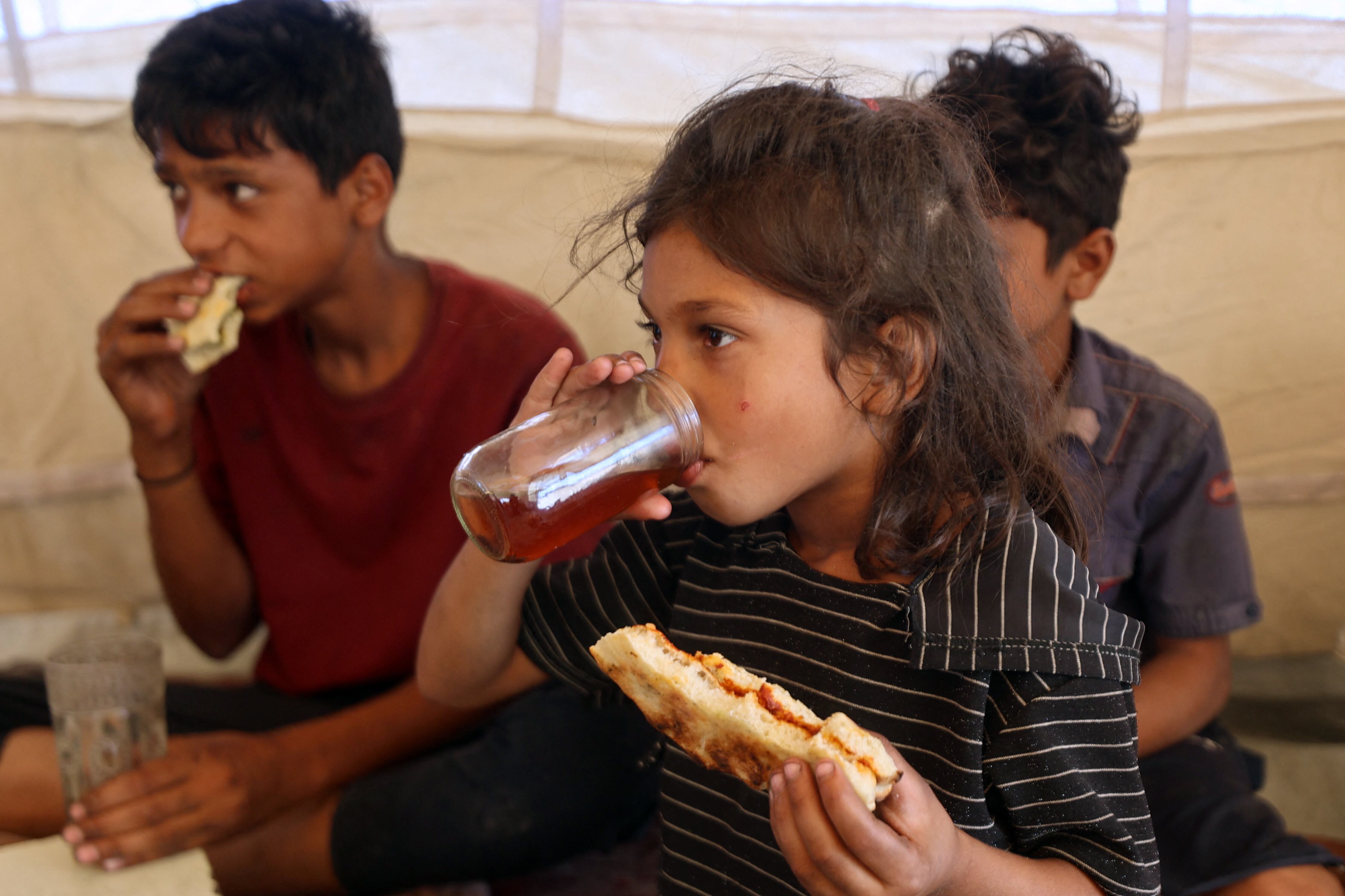 Niños palestinos comen en una tienda de campaña en un campamento temporal para desplazados, instalado en medio de un cementerio en Khan Yunis, al sur de la Franja de Gaza, el 25 de septiembre de 2025, en medio del conflicto entre Israel y el grupo militante palestino Hamás.