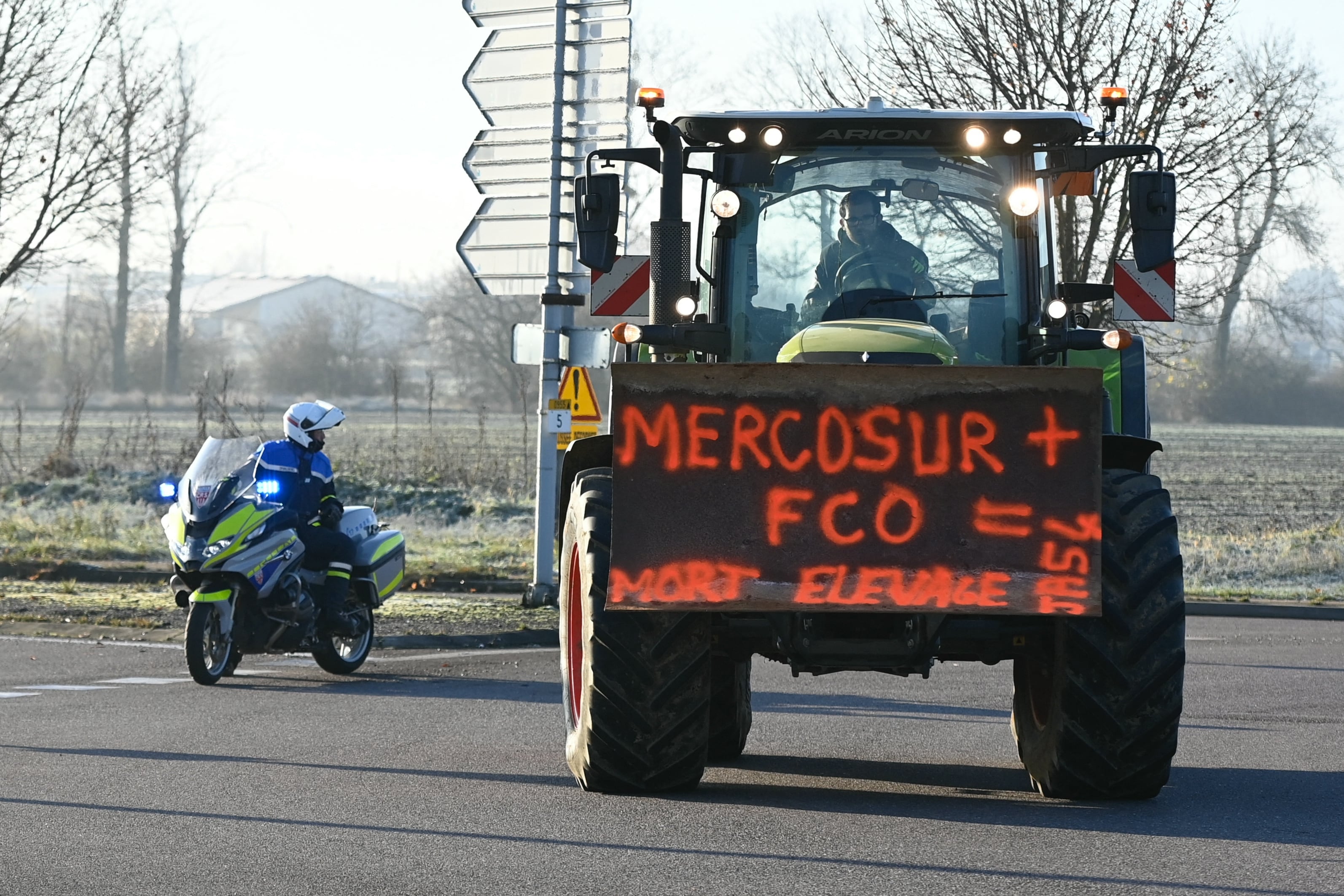 Un tractor con un cartel de protesta contra el acuerdo Mercosur-Unión Europea estacionado en una carretera en Francia, con un policía en motocicleta al fondo.