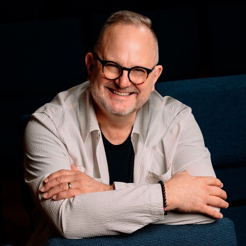 Imagen en primer plano del cantante cristiano Marcos Witt. Usa lentes, una camisa negra y una sobre camisa de color gris claro. Sonríe.