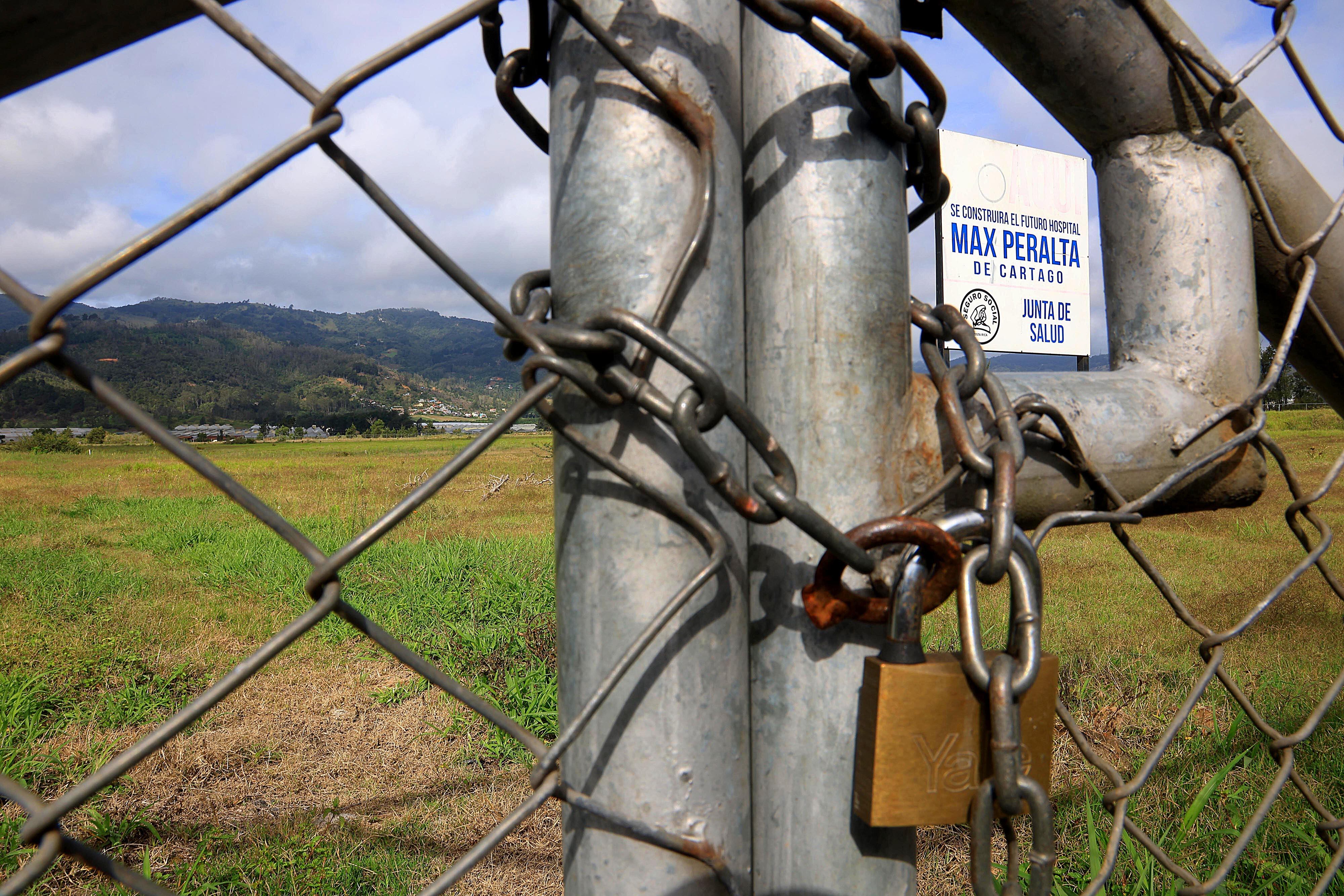 De fondo, el terreno donde se construirá el Hospital de Cartago, con su respectivo rótulo. En primer plano, una malla con un candado cerrado.