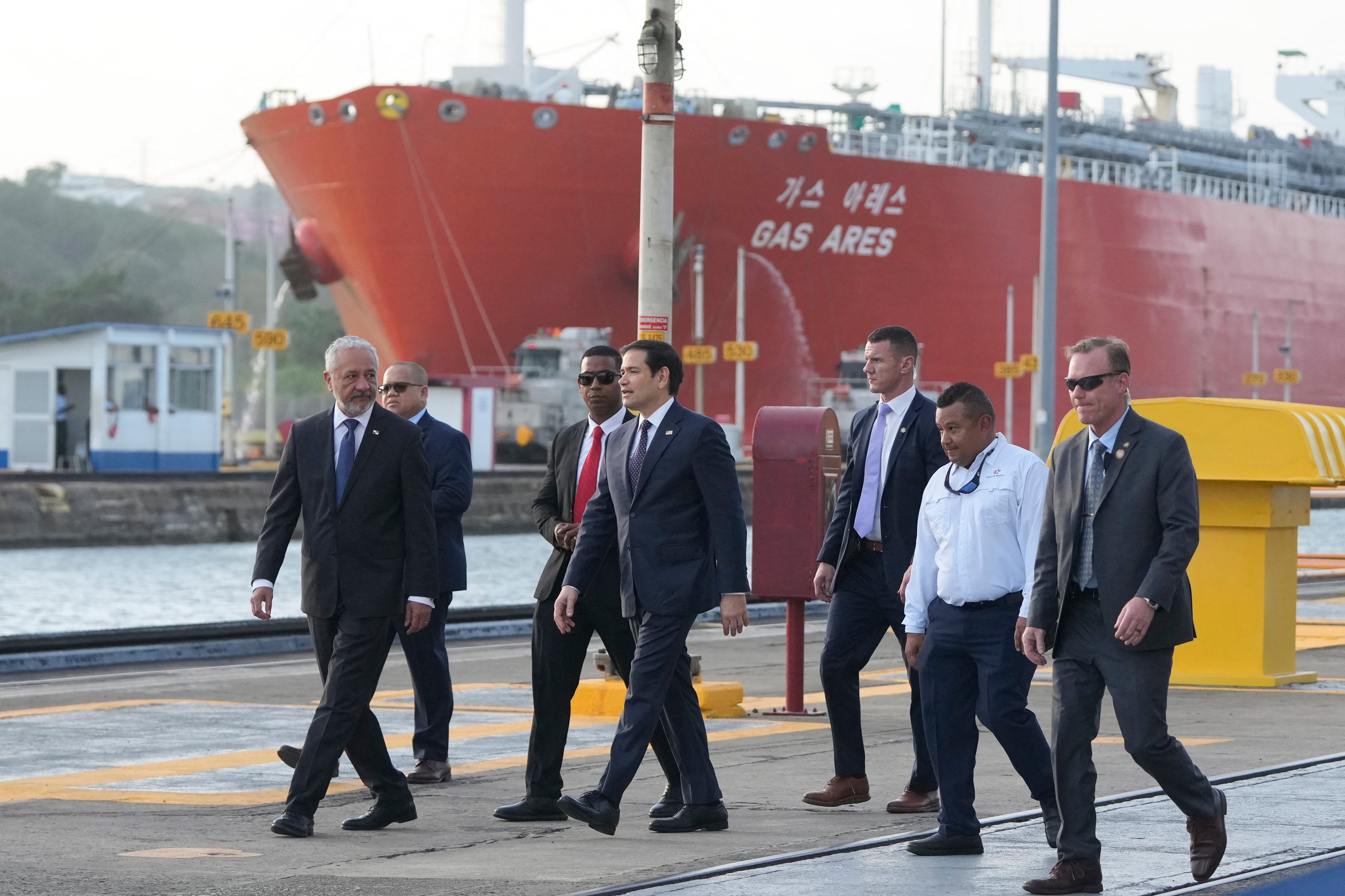 Grupo de hombres en traje entero oscuro caminan en un muelle, junto a un barco de color rojo.