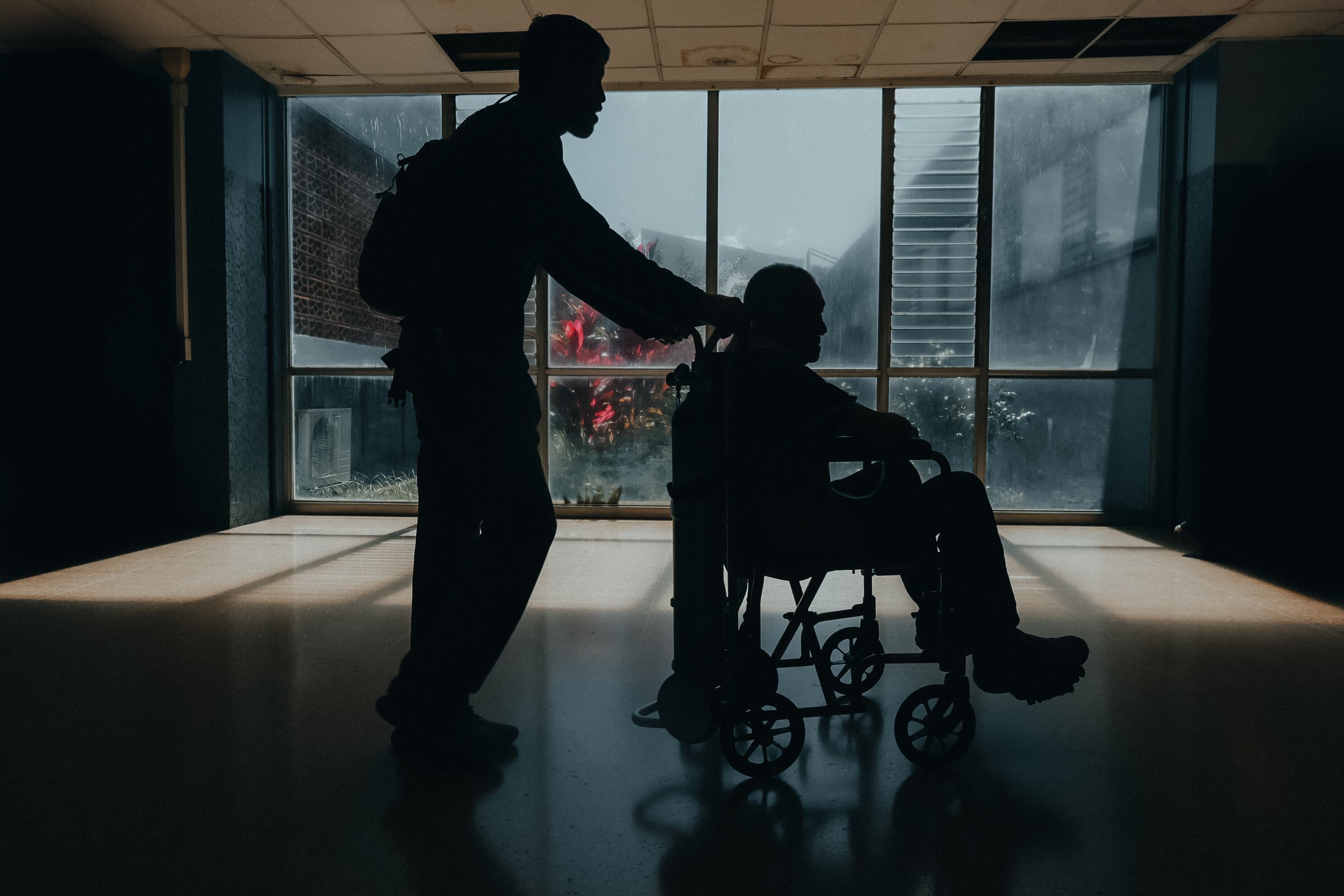 Un adulto mayor es llevado en silla de ruedas por un joven. La imagen está a contraluz. De fondo, se observa una ventana que da a un jardín, en el servicio de consulta externa del Hospital México.
