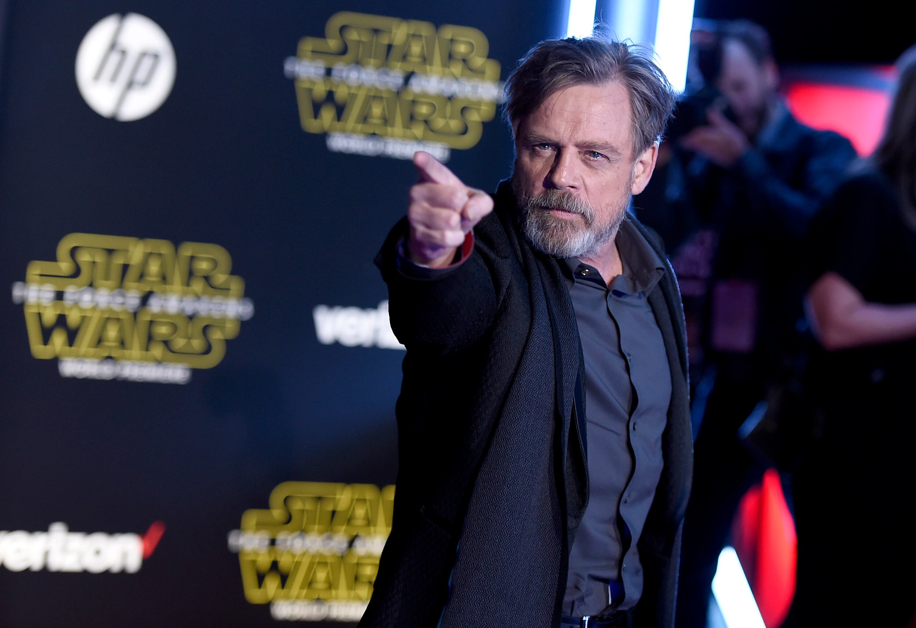 Mark Hamill en una convención sobre Star Wars, se le ve con un traje oscuro y señalando con su mano derecha.