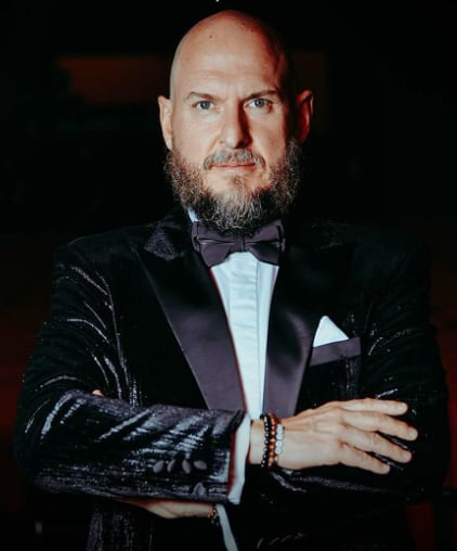 Imagen del actor Andrés Parra en primer plano. Tiene sus brazos cruzados, usa un traje negro, corbatín negro y camisa blanca.