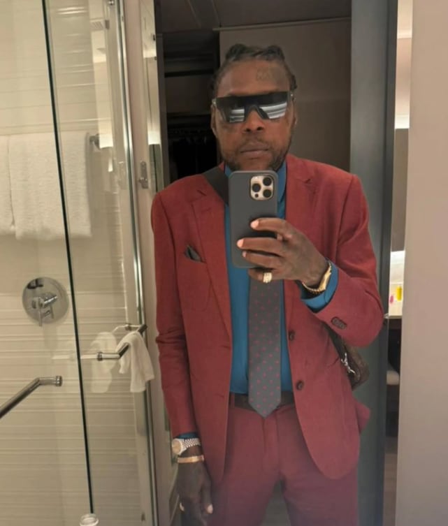 Imagen de Vybz Kartel vestido con un traje de tono rojo, corbata y lentes de sol.