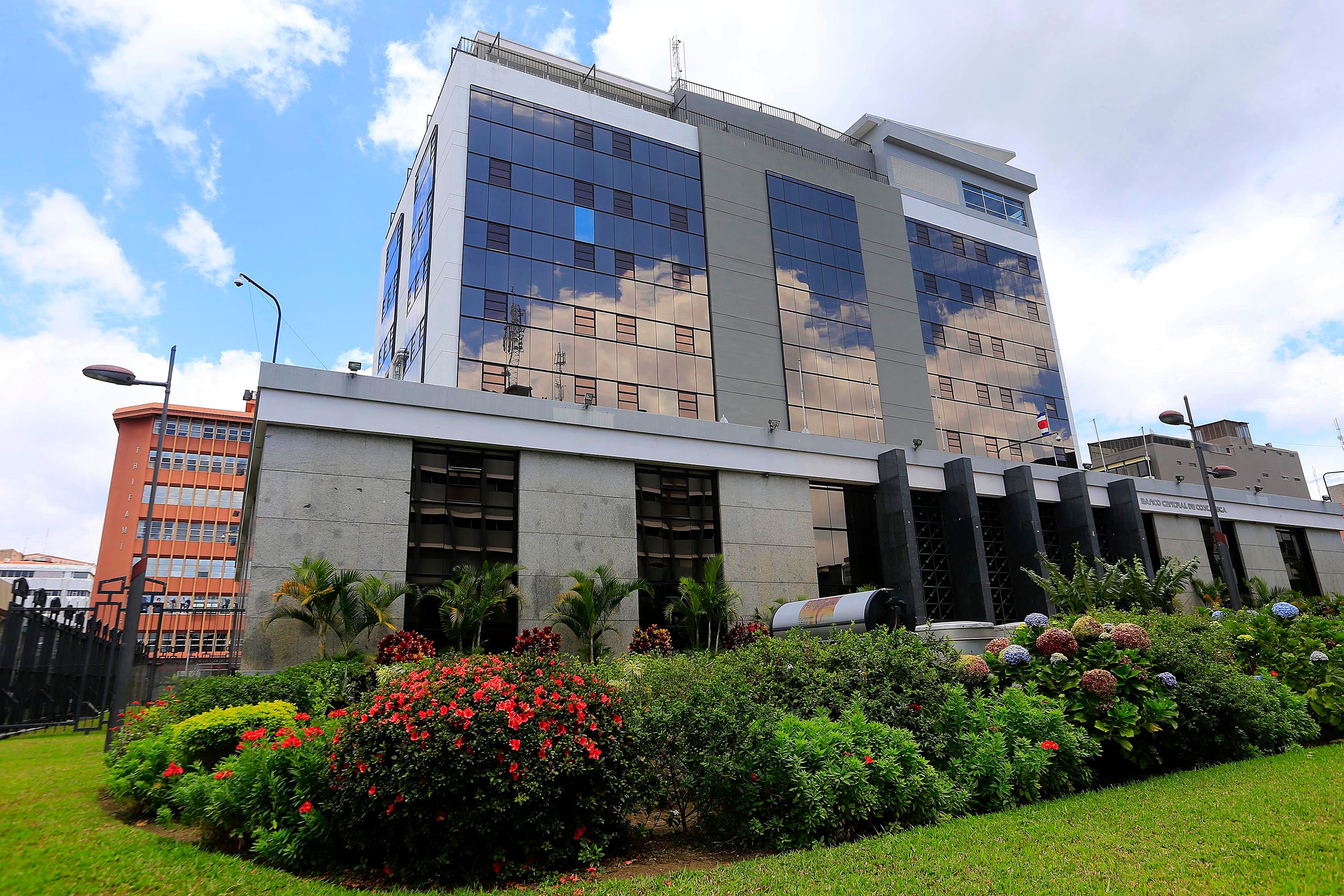 Fachada del edificio del Banco Central de Costa Rica, rodeado de jardines con flores y cielo parcialmente nublado, en San José.