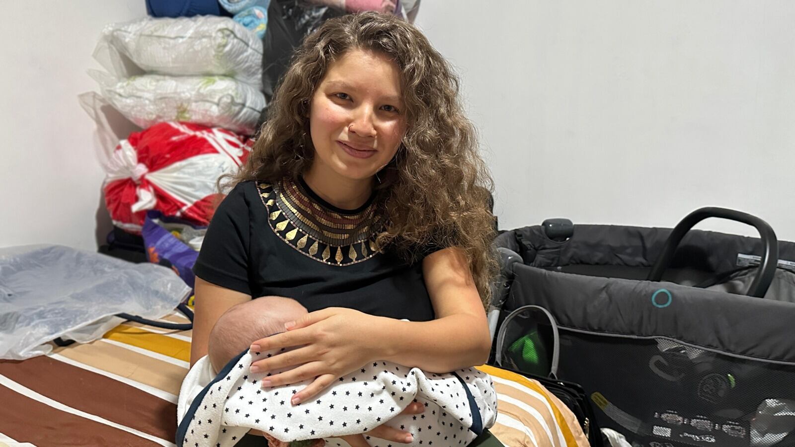 Una mujer joven sostiene a su hijo bebé. En el escenario se ve un coche y ropa de bebé.