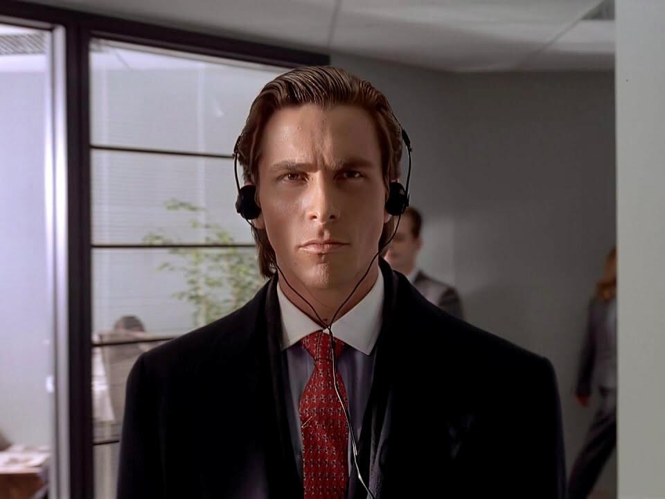 Escena de American Psycho con Patrick Bateman usando audífonos mientras camina por los pasillos de su empresa