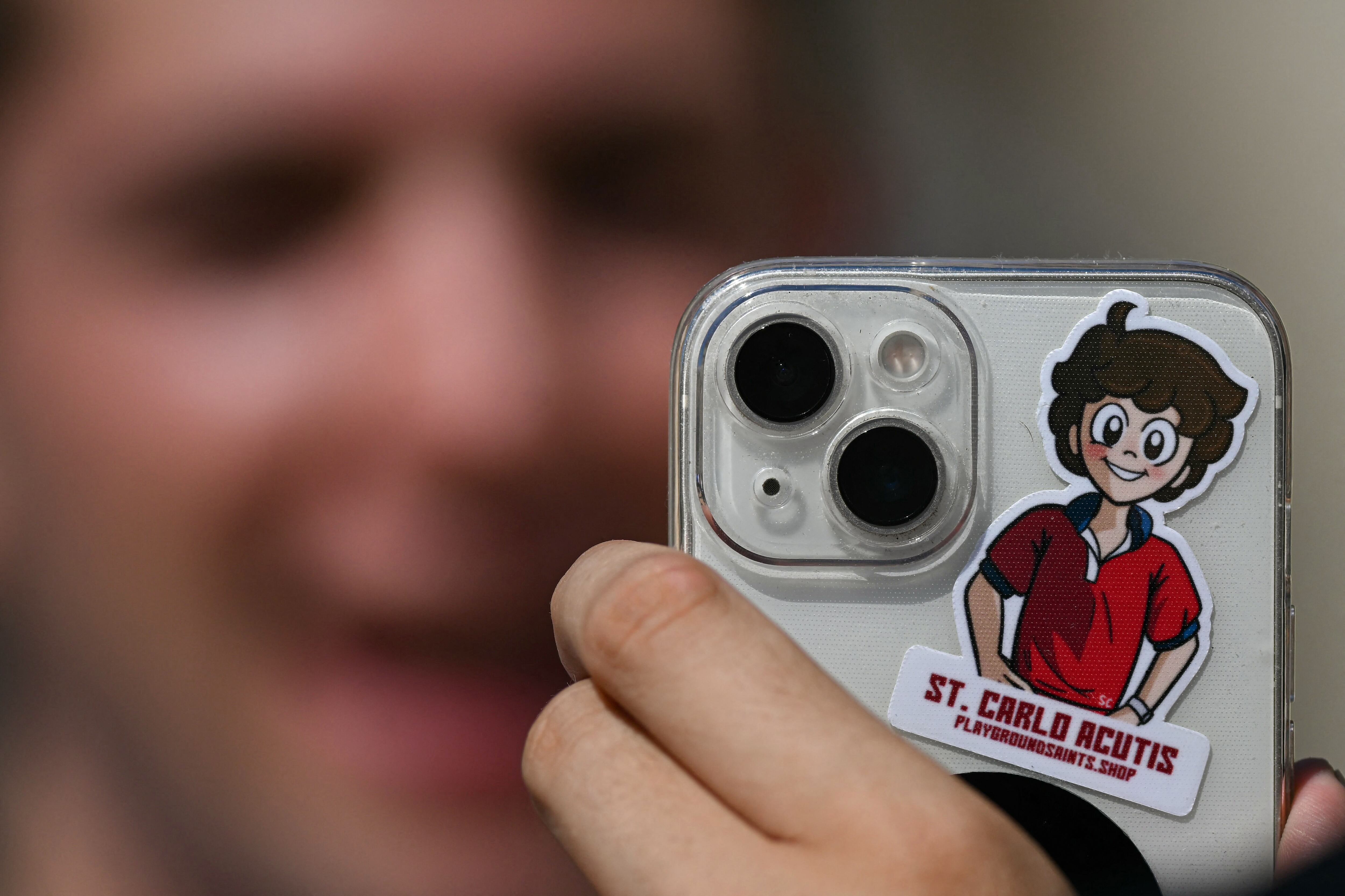 This photograph shows an attendee holding a phone bearing a sticker depicting an image of late Carlo Acutis after a Holy Mass and canonisation of Blessed Carlo Acutis and Pier Giorgio Frassati in St Peter's Square at the Vatican on September 7, 2025. An Italian teenager dubbed "God's Influencer" for his efforts to spread the Catholic faith online will become the first millennial saint Sunday at a canonisation attended by thousands of pilgrims. Computer whiz Carlo Acutis, who died of leukemia in 2006 aged 15, will be raised to sainthood by Pope Leo XIV in a solemn ceremony in St Peter's Square at the Vatican. Italian Pier Giorgio Frassati, a mountaineering enthusiast who died in 1925 and was known for his social and spiritual commitment, will also be made a saint on September 7, 2025. (Photo by Filippo MONTEFORTE / AFP)