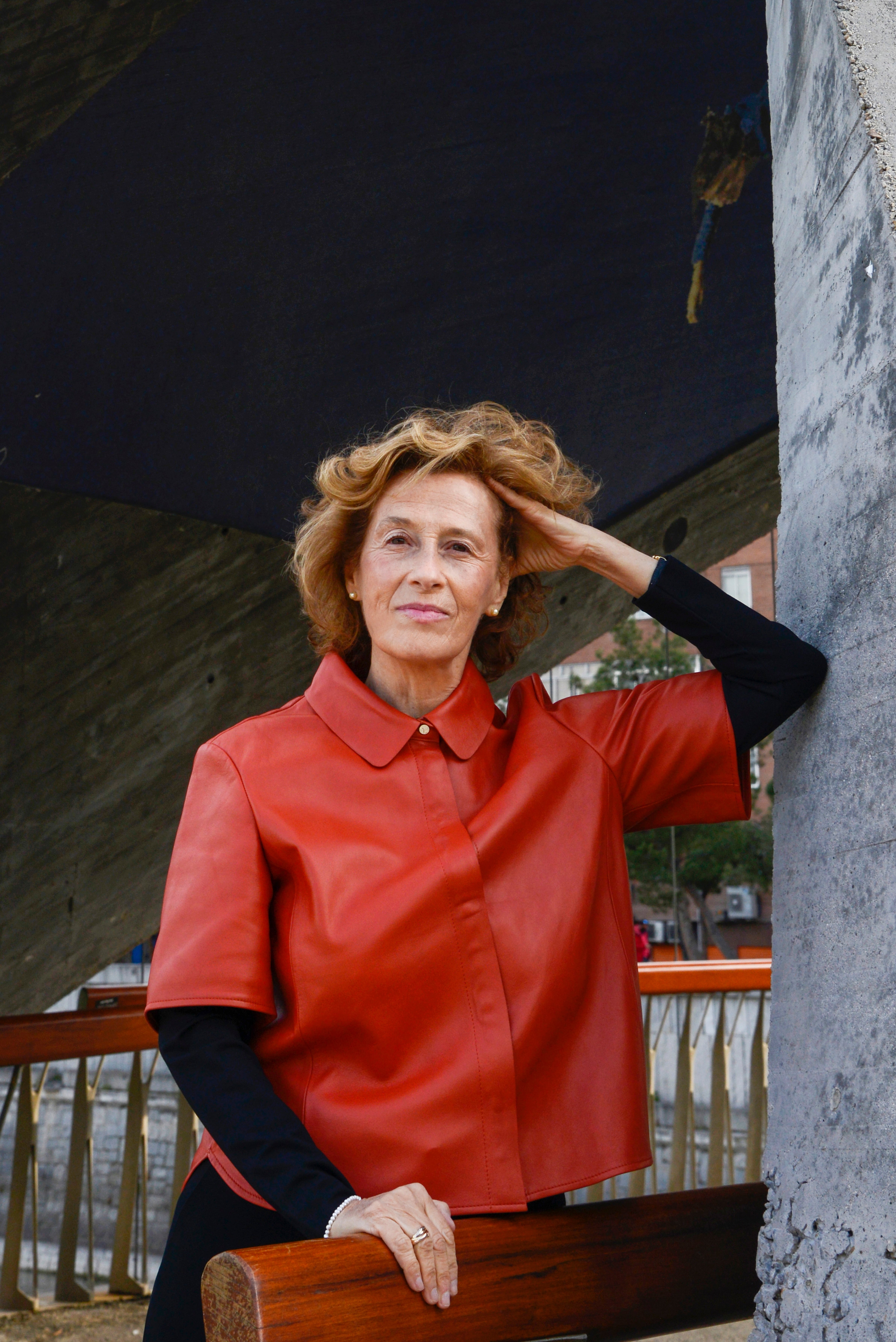 Fotografía de la escritora Julia Navarro, se ve recostada a una pared, usa traje rojo y blusa negra.