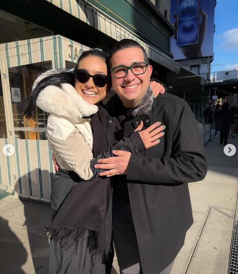 Imagen de la presentadora Nancy Dobles usando un suéter de color crema y lentes para sol abrazando al periodista Elías Alvarado quien luce una chaqueta de color negro.