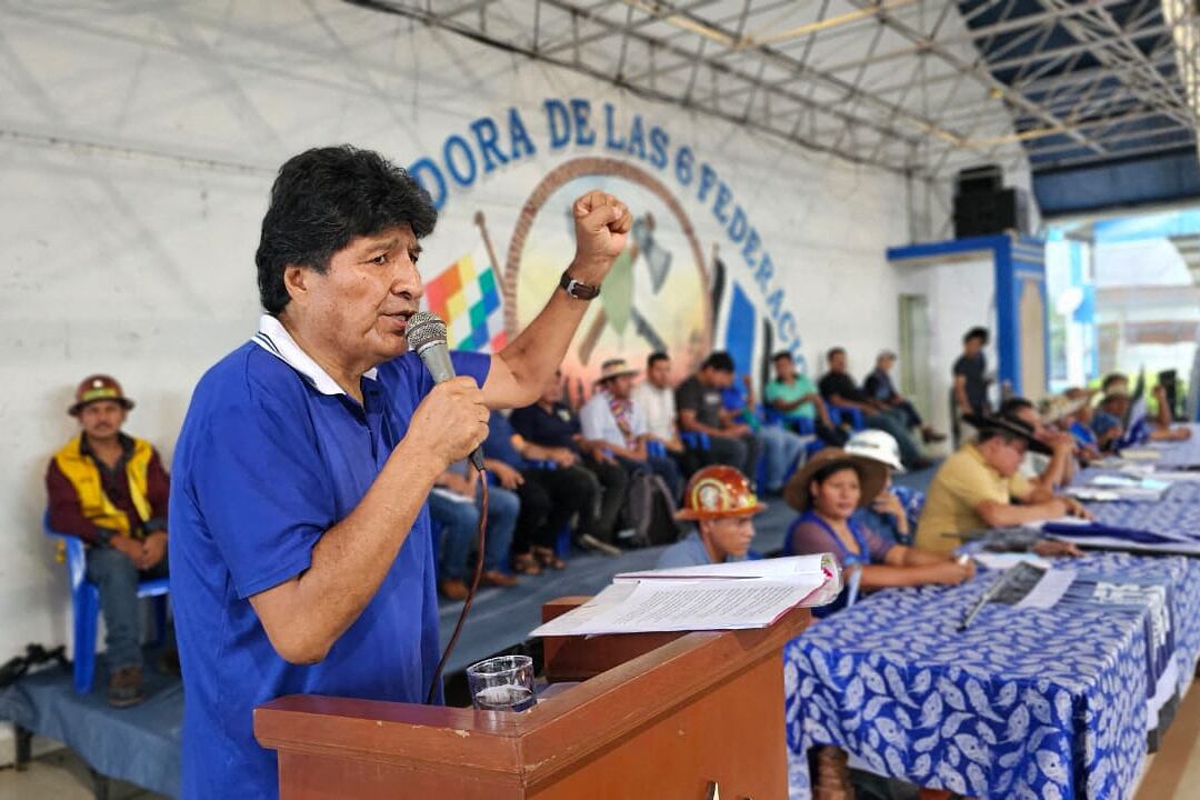 Evo Morales, vestido con camisa azul, habla desde un podio en un evento de la Coordinadora de las 6 Federaciones del Trópico en Bolivia, rodeado de partidarios y con banderas de fondo.
