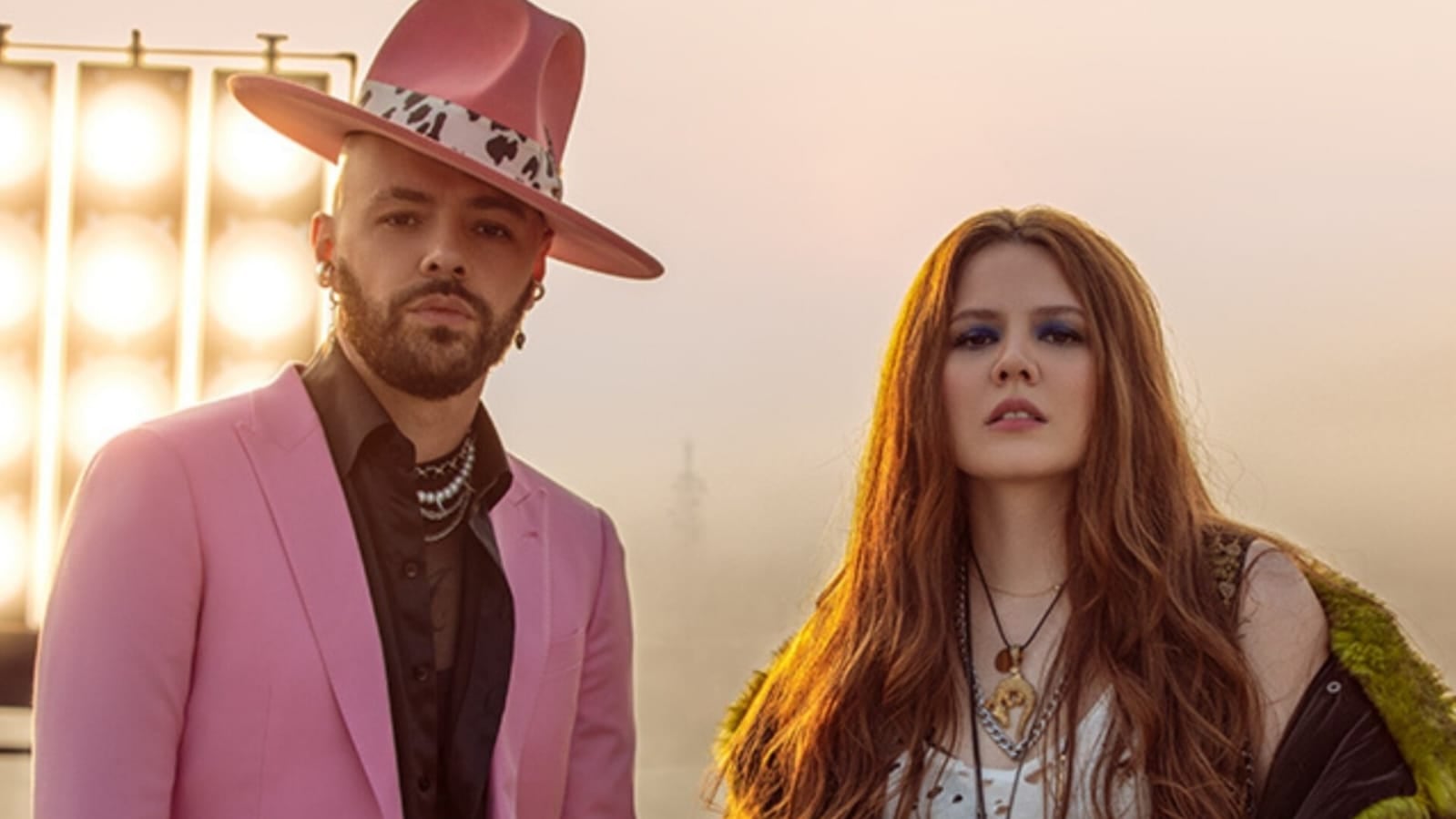 Imagen del dúo Jesse & Joy. Él viste con traje rosado y sombrero y ella con un vestido blanco y sostiene una guitarra en sus manos.