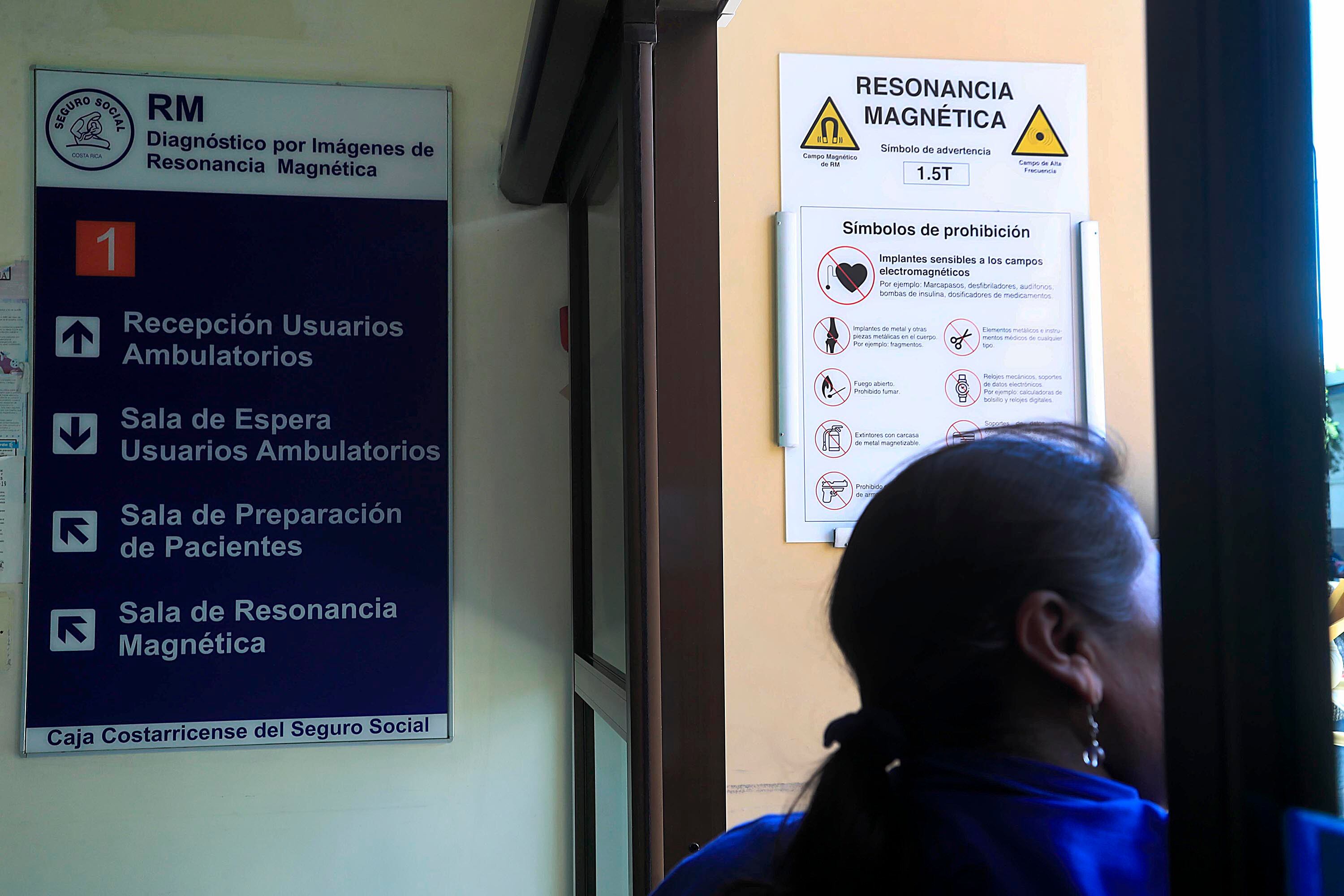 Rótulos en un hospital que indican Servicio de Resonancia Magnética. A contraluz, el perfil de una mujer con blusa azul.